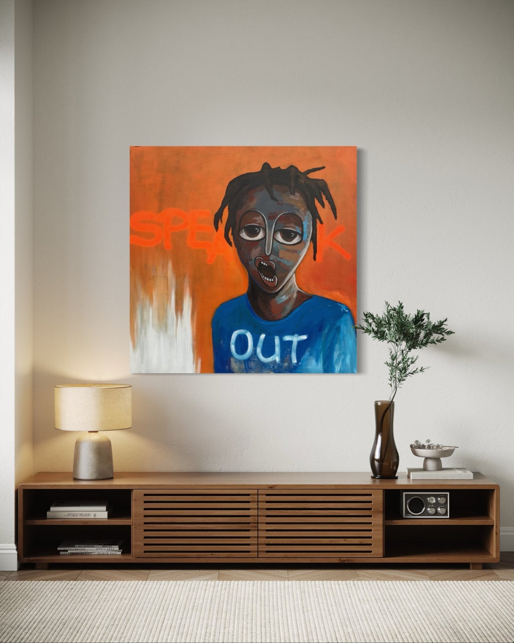 A modern living room with a wooden media console, lamp, vase, and books features "Speak Out," a colorful hand-signed painting by Achike Anayo with an expressive portrait and orange background—included is a certificate of authenticity.