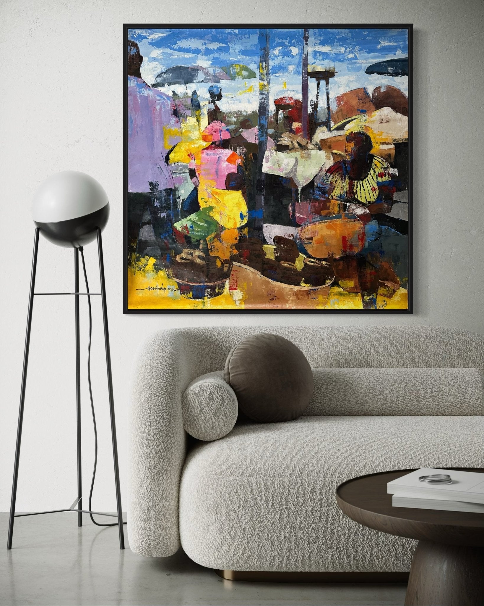 A cozy living room with a textured white sofa, round brown pillow, modern floor lamp, and "Stockfish," a colourful abstract market scene painting by Olasunkanmi Oyelusi—hand-signed and with certificate—adorning the white wall.