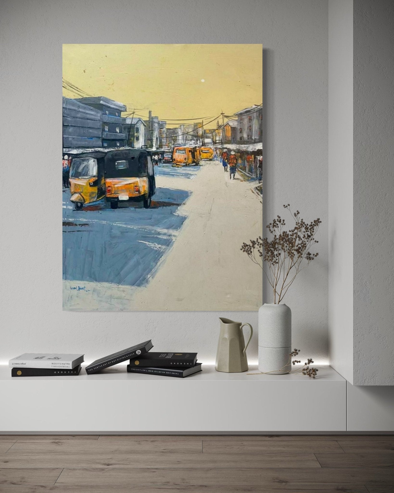 A modern, minimalist room features "Streets Of Lagos" by Lucky Isaiah—a hand-signed painting of a vibrant yellow city street with tuk-tuks and people—displayed above a low white shelf holding books, a ceramic jug, and a vase with dried branches.