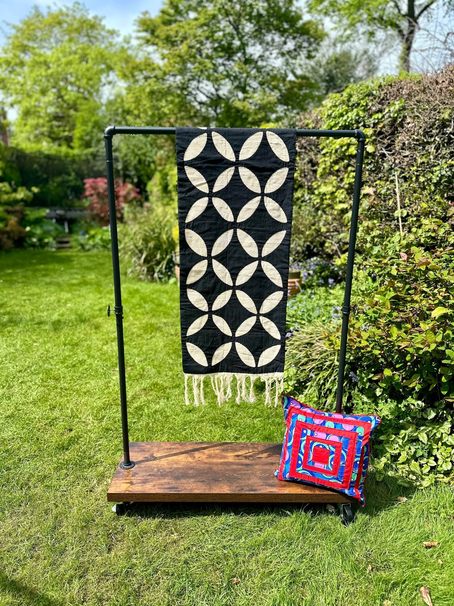 A Table Runner Black by bibianna.co.uk with a white geometric pattern hangs on a metal rack outdoors, while a colorful square pillow featuring red, blue, and white designs rests nearby among green grass and foliage.