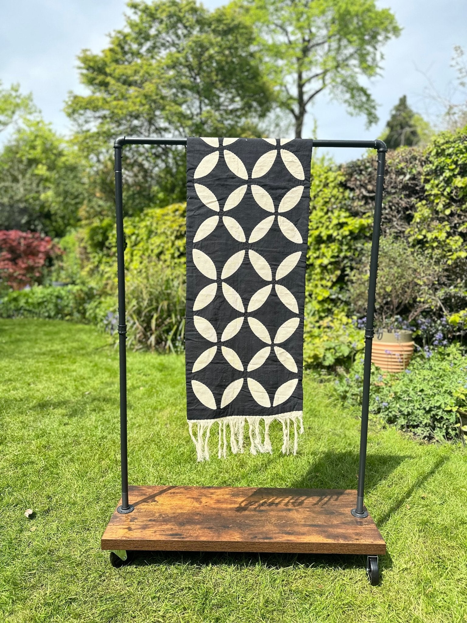 The Table Runner Black by bibianna.co.uk, featuring a bold geometric pattern in black and cream with fringe, is displayed on a metal rack with a wooden base, set outdoors on lush green grass with trees and plants in the background.
