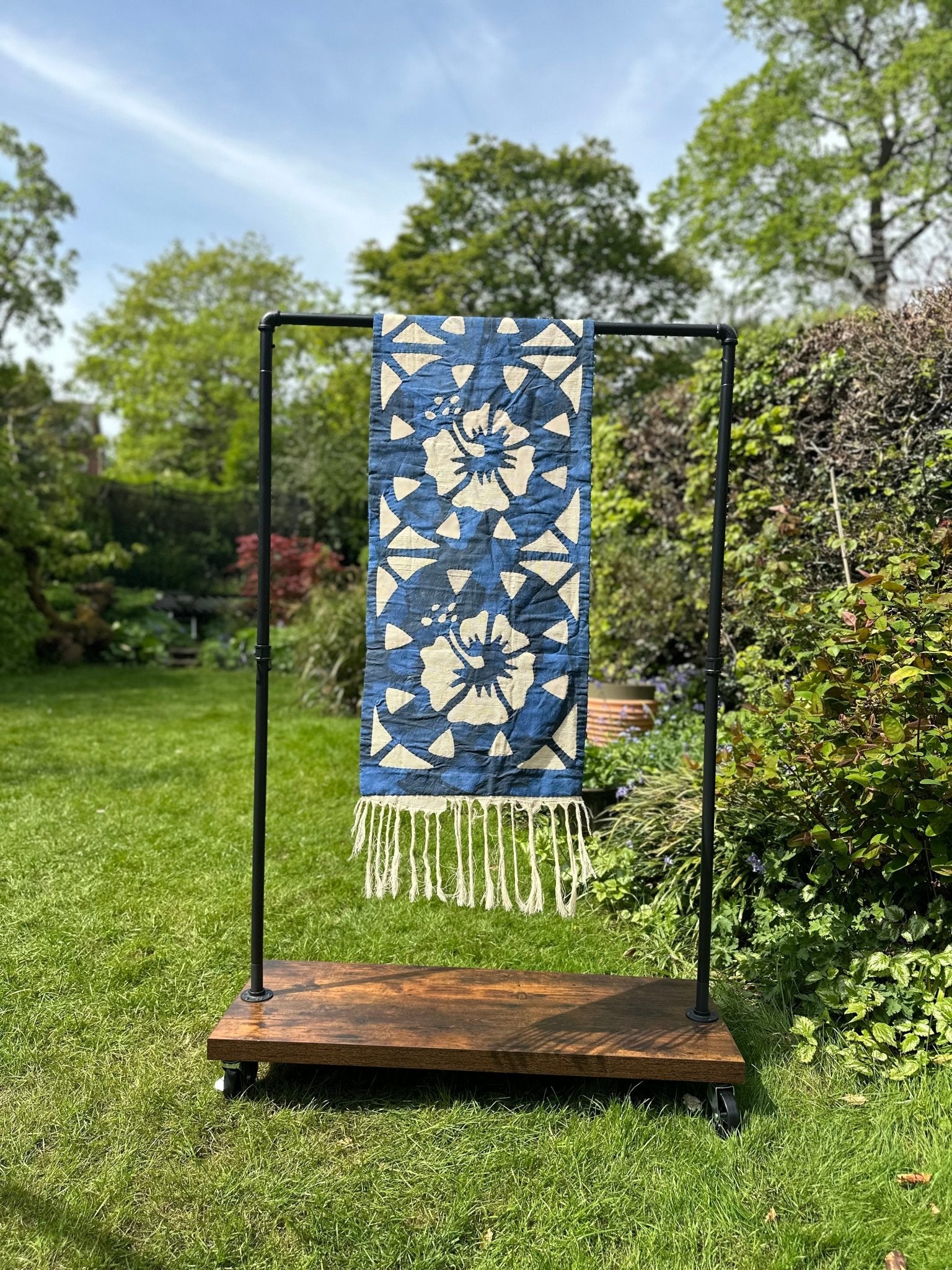 A Table Runners Blue by bibianna.co.uk, featuring blue and white patterns with fringe, is displayed on a black metal and wood stand in a lush green garden surrounded by trees and plants.