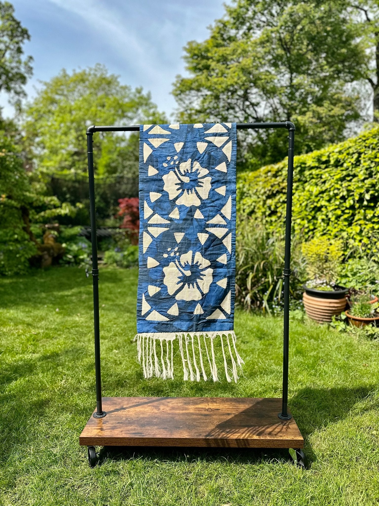 The Table Runners Blue by bibianna.co.uk, featuring a blue and white pattern with fringes, hangs on a black metal rack with a wooden base, displayed outdoors on green grass amid trees and bushes.