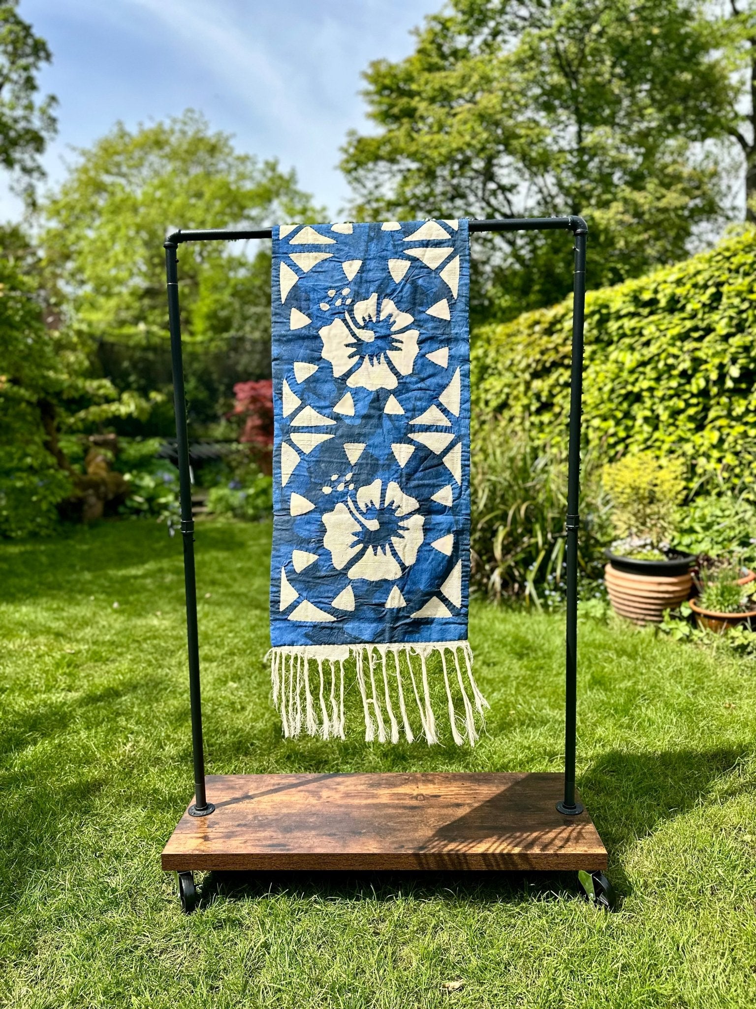 The Table Runners Blue from bibianna.co.uk features a blue and white pattern with fringe, ideal for African-inspired decor. Displayed on a black metal rack with a wooden base, it enhances any setting with natural greenery and flowers in the background.
