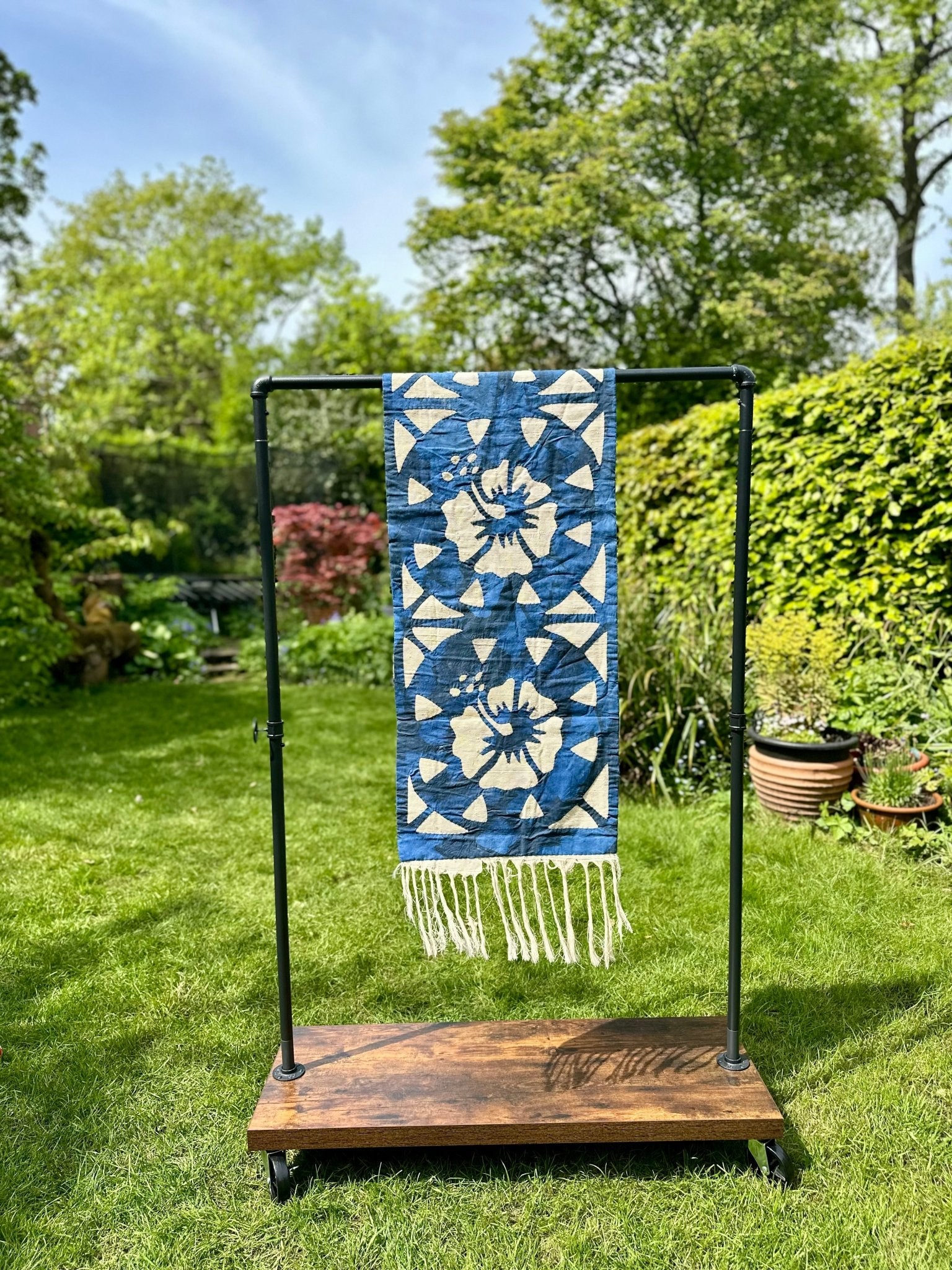 The Table Runners Blue by bibianna.co.uk, featuring a blue and white pattern, is displayed as a table runner on a black metal rack with a wooden base, set outdoors against green trees and plants.