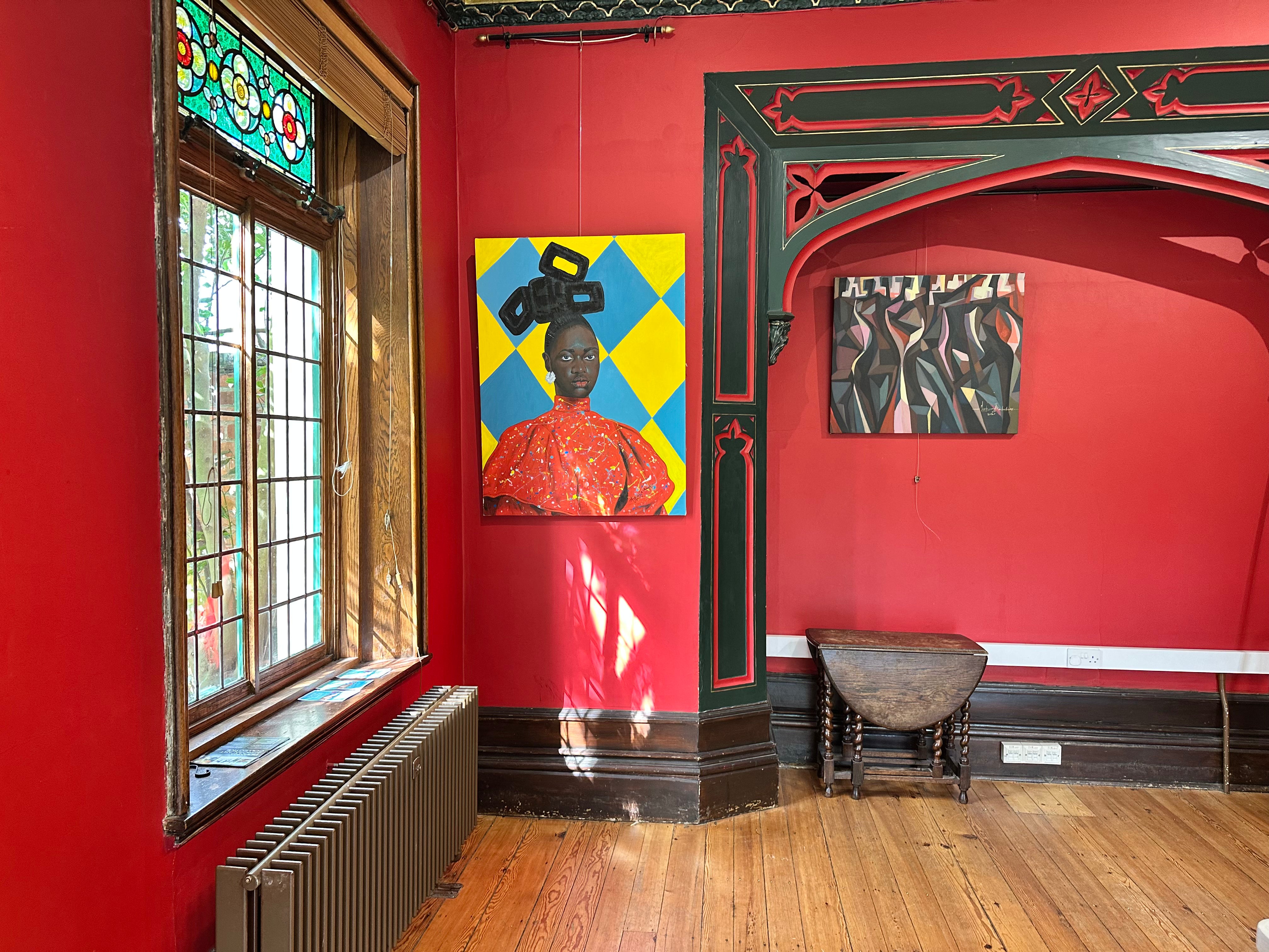 A vibrant red-walled room features Barry Ikechukwu's 'The Red Man' (78x78 cm), depicting abstract human forms in red with a yellow and blue diamond-patterned background, alongside a cubist artwork. Sunlight filters through stained glass.