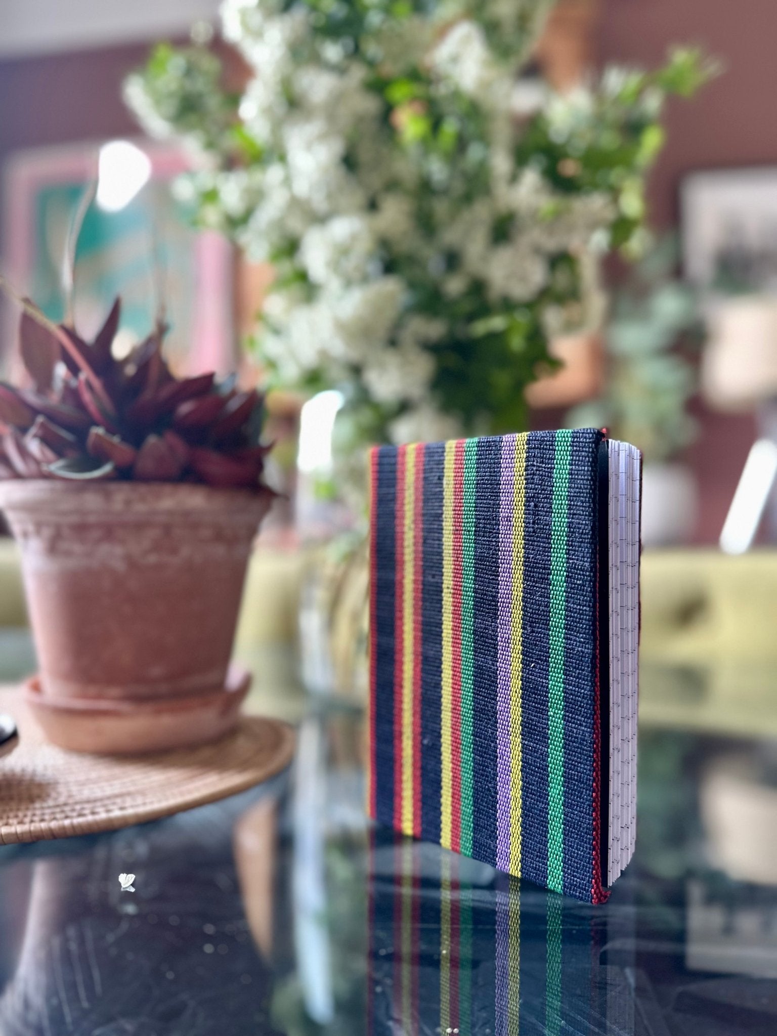 The Ashoke Notebooks A5 & A6 by bibianna.co.uk, featuring a colorful stripe design inspired by Nigerian Ashoke fabric, stand upright on a glass table beside a potted succulent. In the background are white flowers and framed art.