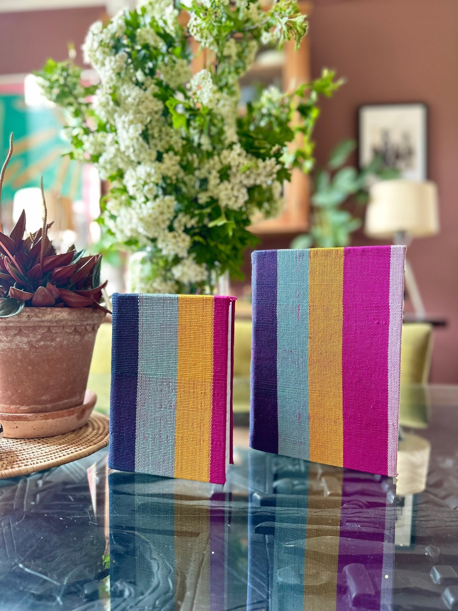 The Ashoke Notebooks A5 & A6 by bibianna.co.uk, one large and one small with colorful textured covers, stand upright on a glass table, backed by a potted succulent and white flowers in a softly blurred indoor setting.