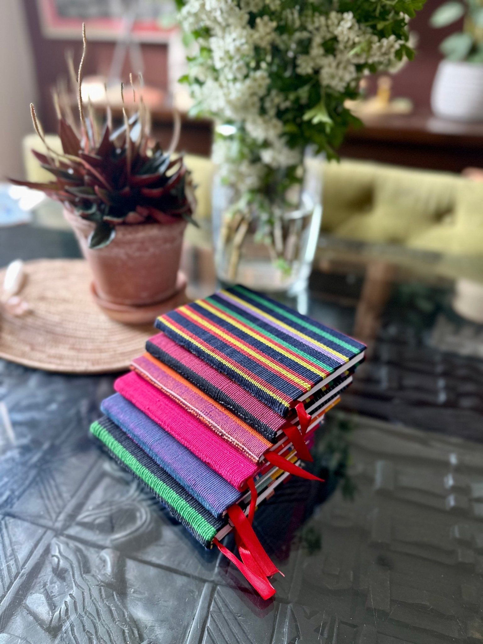 A stack of bibianna.co.uk Ashoke Notebooks A5 & A6, covered in vibrant Nigerian Ashoke fabric with red ribbons, rests on a glass table beside a potted plant, with a vase of white flowers adding to the cozy setting.