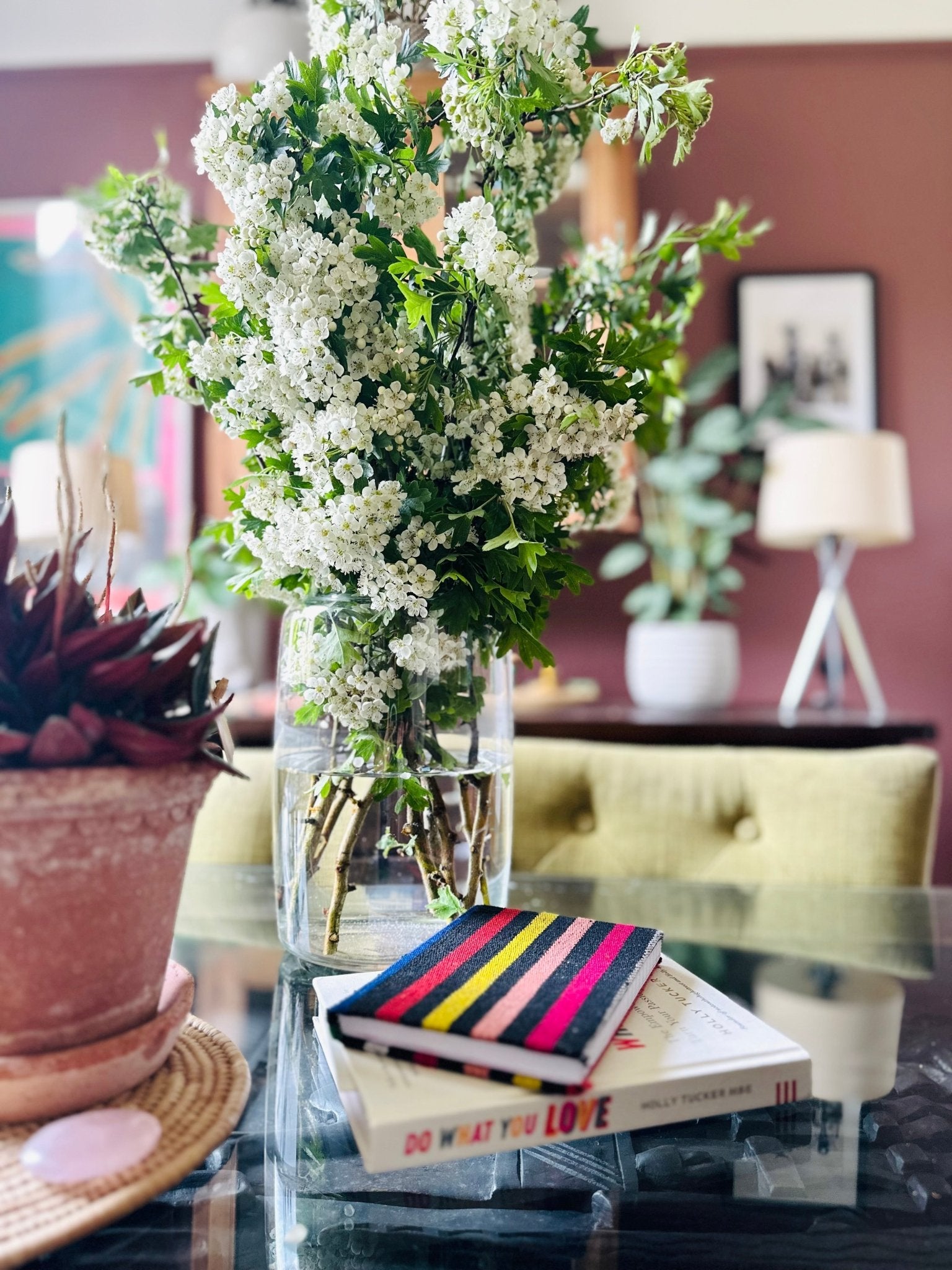 A glass vase of white flowers sits on a glass table beside a potted plant, with two books and bibianna.co.uk's Ashoke Notebooks A5 & A6, inspired by Nigerian Ashoke fabric, stacked nearby; a cozy living room is in the background.