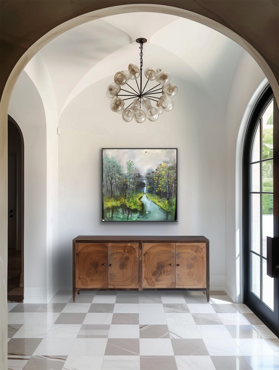 A modern entryway with a checkered tile floor and wooden sideboard features "The Great Outdoors" by Oyebisi Elijah—hand-signed artwork—beneath an arched ceiling and round chandelier, with light streaming through a large glass door.