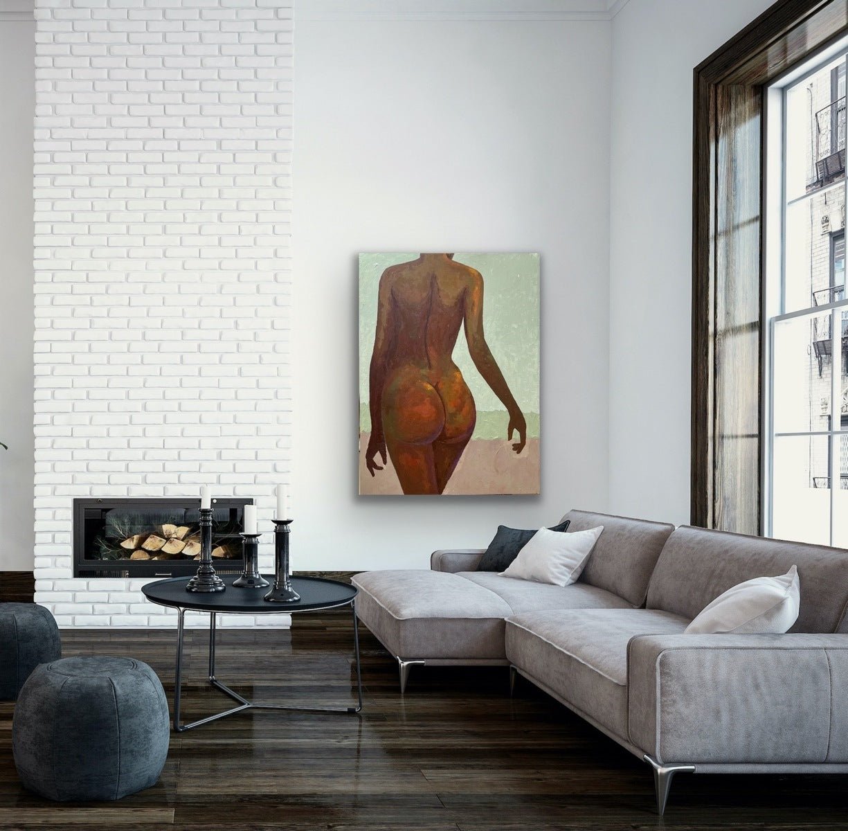 A modern living room features a gray sectional, round black coffee table, two ottomans, and “The Temple” Tasteful Painting Nude by Faith O Sidneu displayed above the fireplace on a white brick wall. Large windows fill the space with natural light.