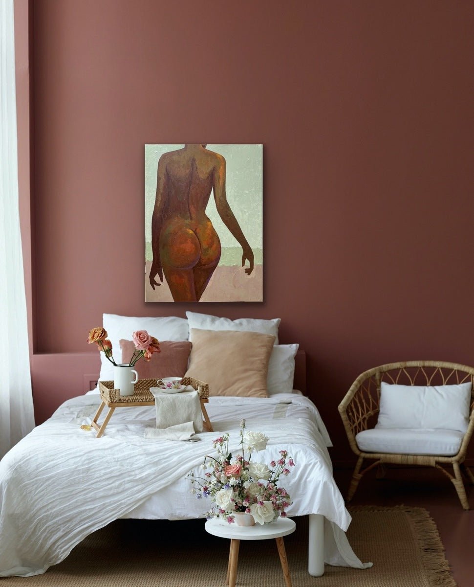 A cozy bedroom with red-brown walls, a white bed with beige pillows, a wicker chair, and a tray set. Above the bed hangs "The Temple" Tasteful Painting Nude by Faith O Sidneu. A vase of flowers rests on a small table nearby.
