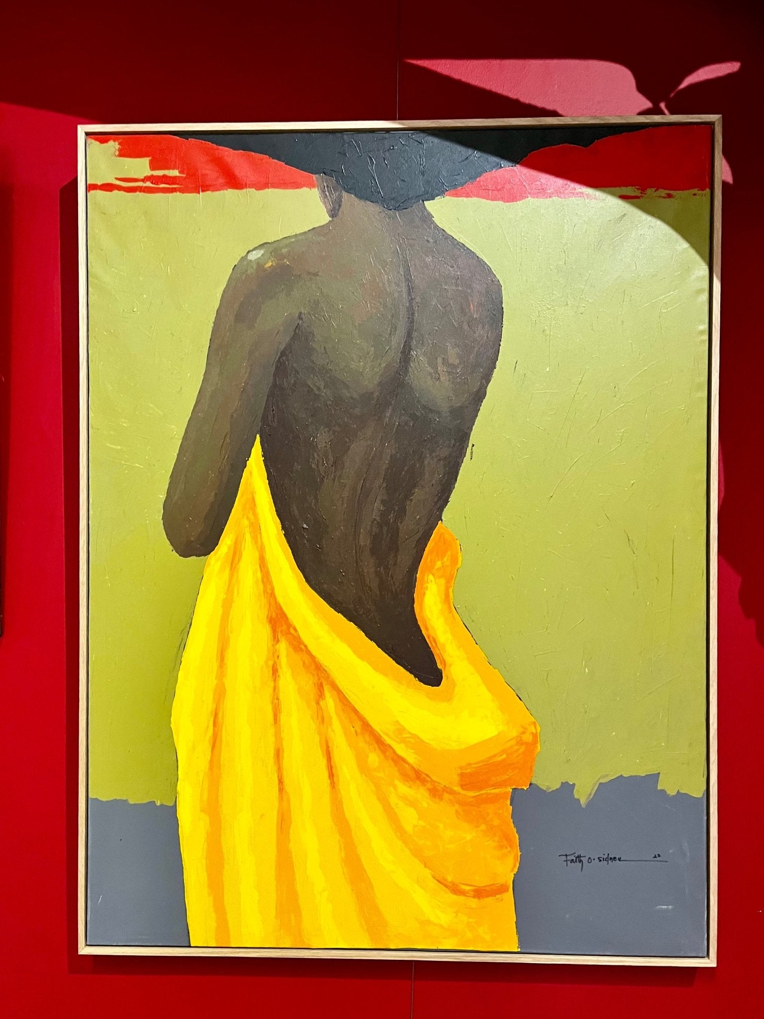 “The Yellow Fabric” by Faith O Sidneu shows a figure with a bare back in bright yellow cloth, against a green and red backdrop. Bold brushstrokes enliven this acrylic on canvas as the person’s head is turned away.