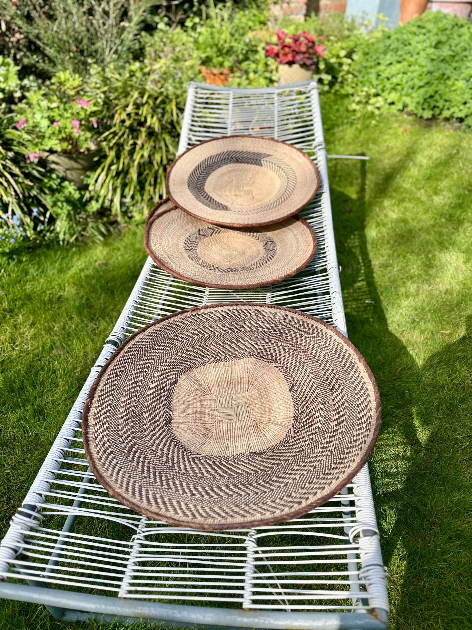 Three Tonga / Binga Basket Zimbabwean Basket Wall Art (C) by bibianna.co.uk are shown on a white metal bench in a sunny garden, making them perfect as textured wall art for your home décor.