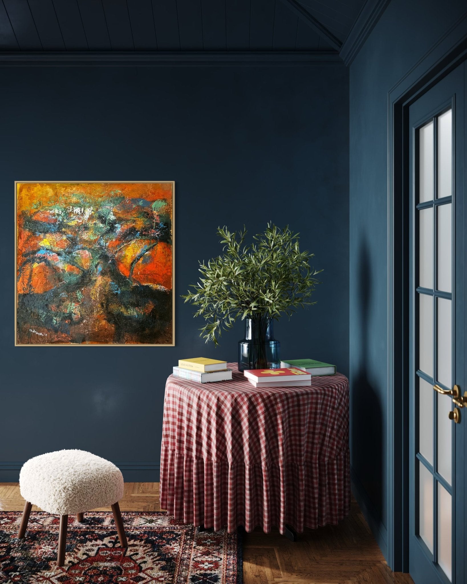 A cozy nook with dark blue walls features “Tree Of Life”—an abstract orange painting hand-signed by Bibianna African Art Collective—hanging above a round table with a red checked cloth, books, a vase of greenery, fuzzy stool, and patterned rug on wood floors.