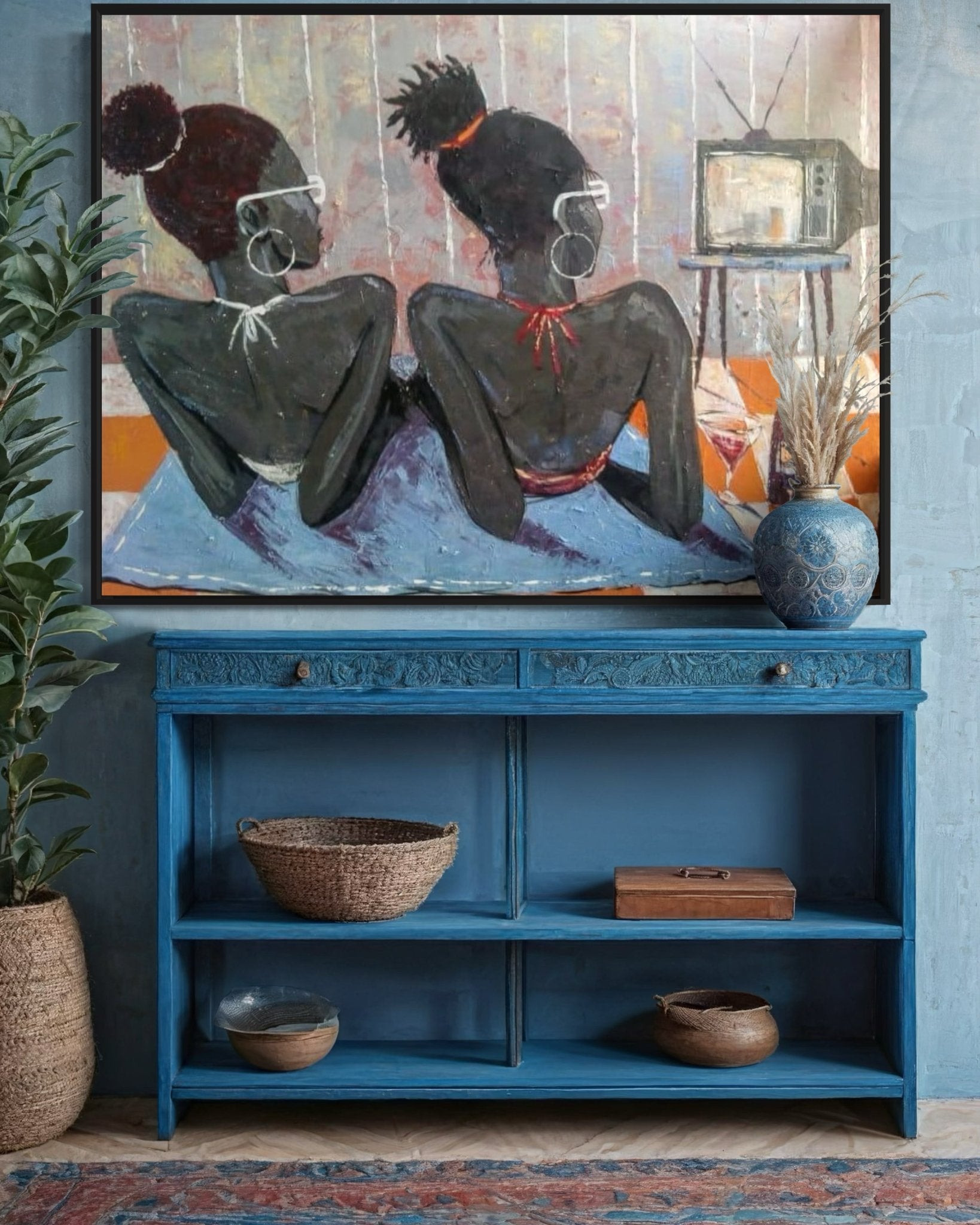 Lucky Isaiah's "Tween Vacation," a framed, hand-signed artwork of two women lying side by side watching TV, is displayed above a blue console with baskets and vases. A certificate of authenticity is included for this cozy, artistic piece.