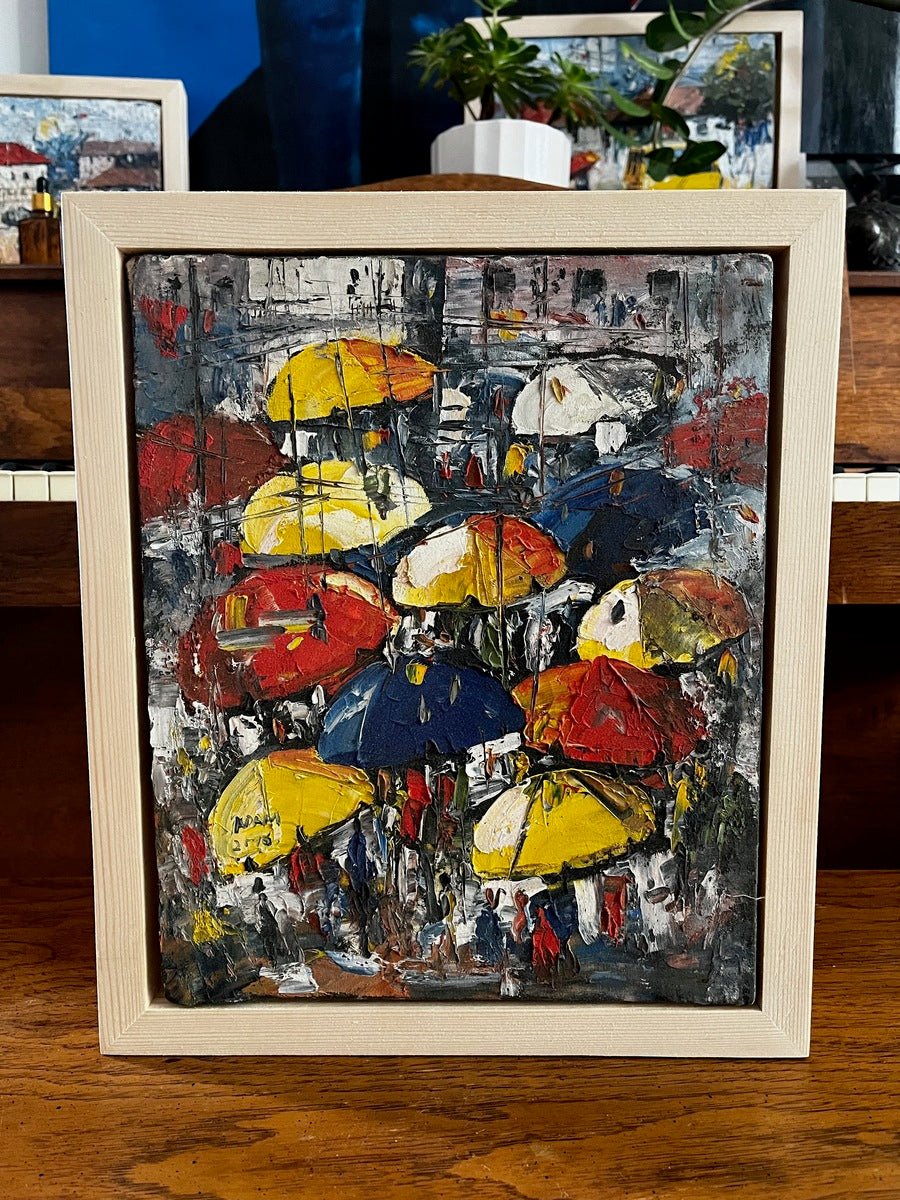 The "Umbrellas in The Sun. Street Market" by Adam is a framed, heavily textured miniature acrylic on canvas painting showing a vibrant crowd with colorful umbrellas, displayed on a wooden surface with plants and art nearby.