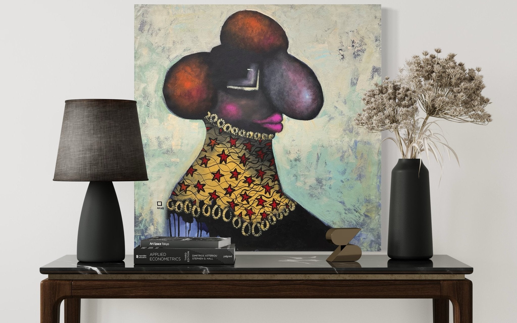 A console table with a black lamp, books, and a vase of dried flowers sits below "Untitled" Contemporary Painting by Prince Obasi—an acrylic on canvas portrait featuring bold colors and exaggerated features.