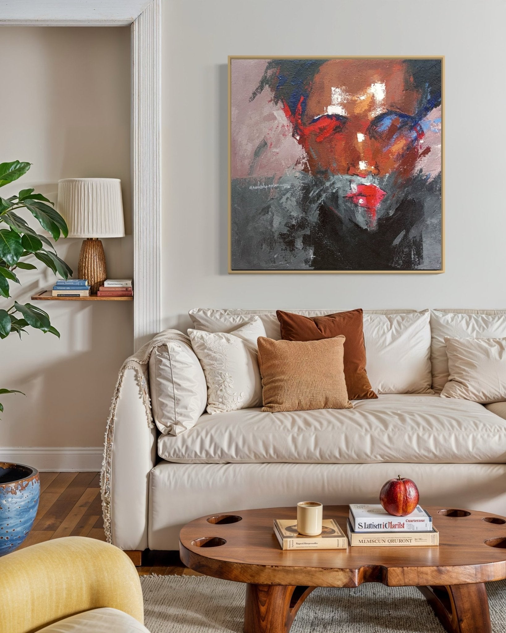 A cozy living room with a cream sofa, brown and beige cushions, a wooden coffee table with books and a candle, and "Where I Am" (93x93cm) by Olasunkanmi Oyelusi displayed above the sofa.