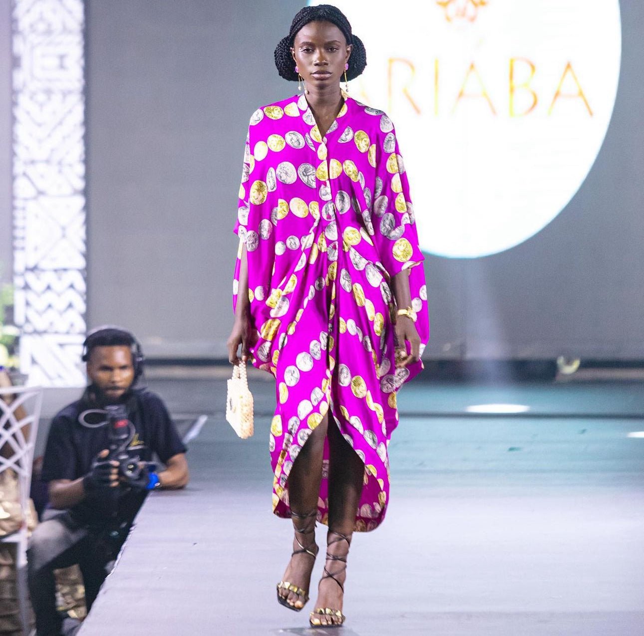 A model walks the runway in a vibrant pink and yellow Zoya Drapped Dress by bibianna.co.uk, featuring a high slit. She carries a small handbag, wears strappy sandals, while a photographer is visible near the lit display in the background.