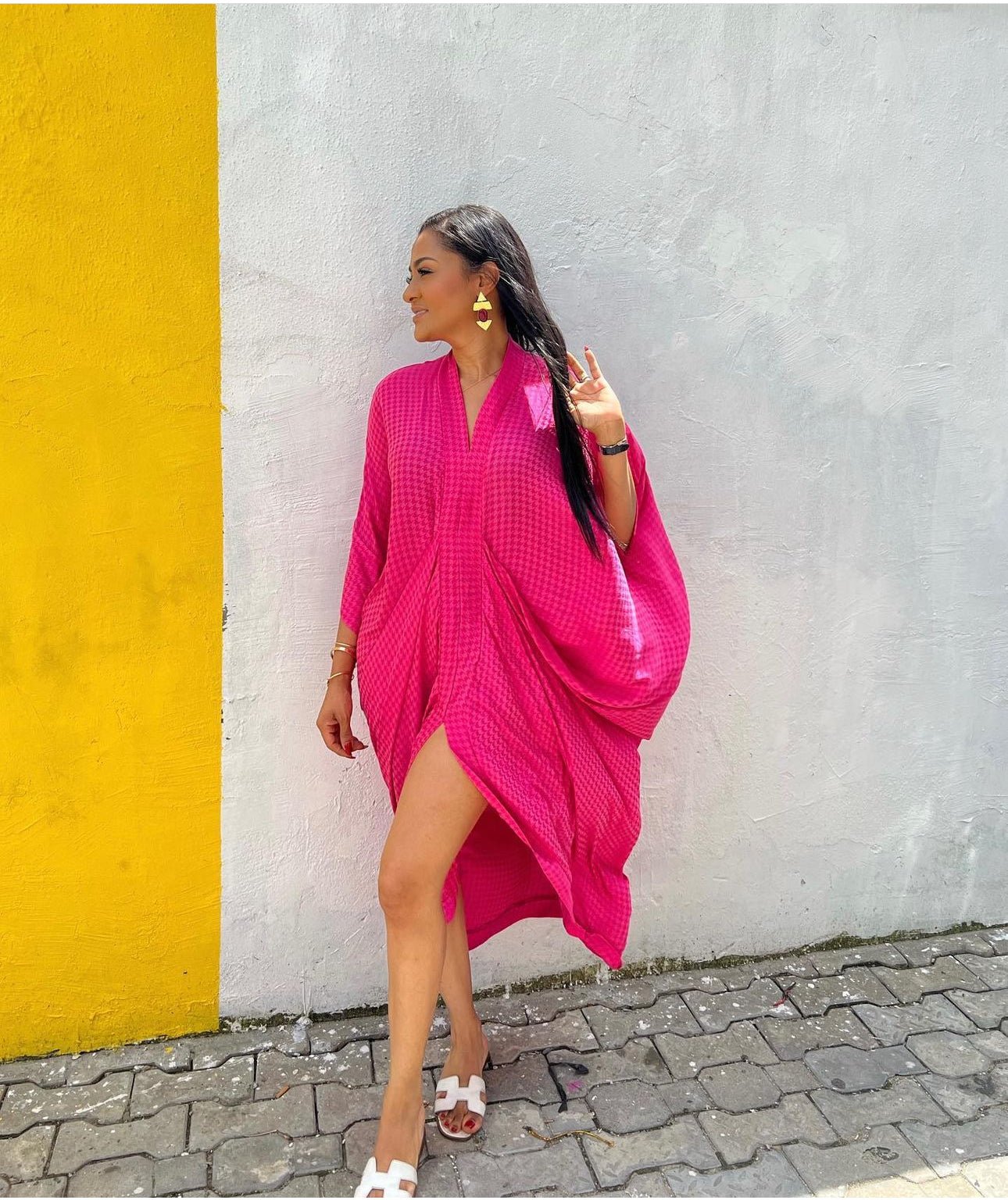 A woman wears the Zoya Drapped Dress from bibianna.co.uk, posing with one leg forward and showing a peace sign on a cobblestone sidewalk in front of a white wall with a yellow stripe. She pairs it with white sandals and gold earrings.