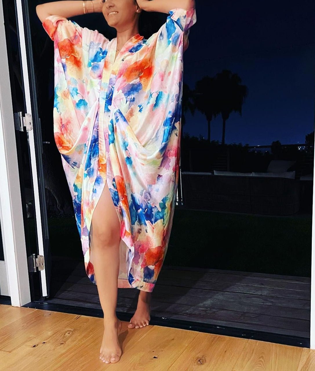 Barefoot in a doorway at night, a woman wears the vibrant, loose Zoya Drapped Dress by bibianna.co.uk with one leg revealed. Silhouetted trees and darkness outside highlight the bestseller’s elegance in this tranquil scene.