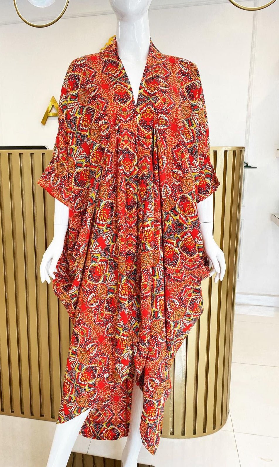 A mannequin displays the Zoya Drapped Dress by bibianna.co.uk—a loose, red-orange midi dress with wide sleeves and draped design—set in a modern indoor space with gold vertical panels in the background.