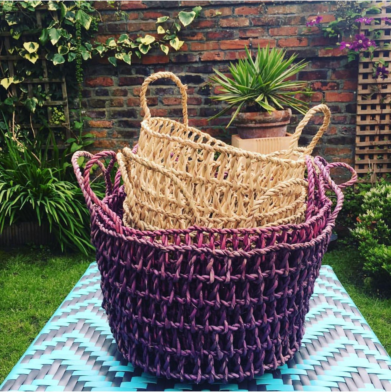 Three woven baskets, two beige and one purple, are nested together on a blue patterned table in a garden with green plants, a brick wall, and potted plants in the background.