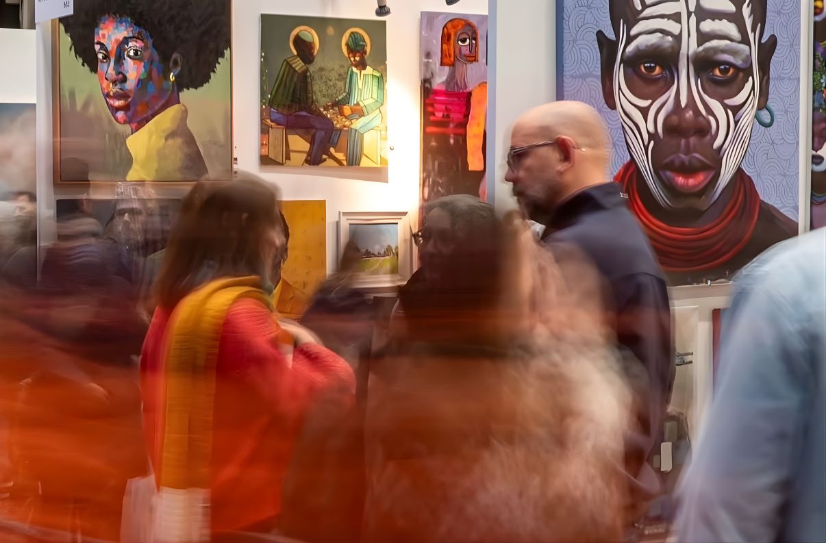 A group of people converse in front of vibrant, colorful portrait paintings at an art gallery; some figures are blurred, capturing movement and a lively atmosphere.