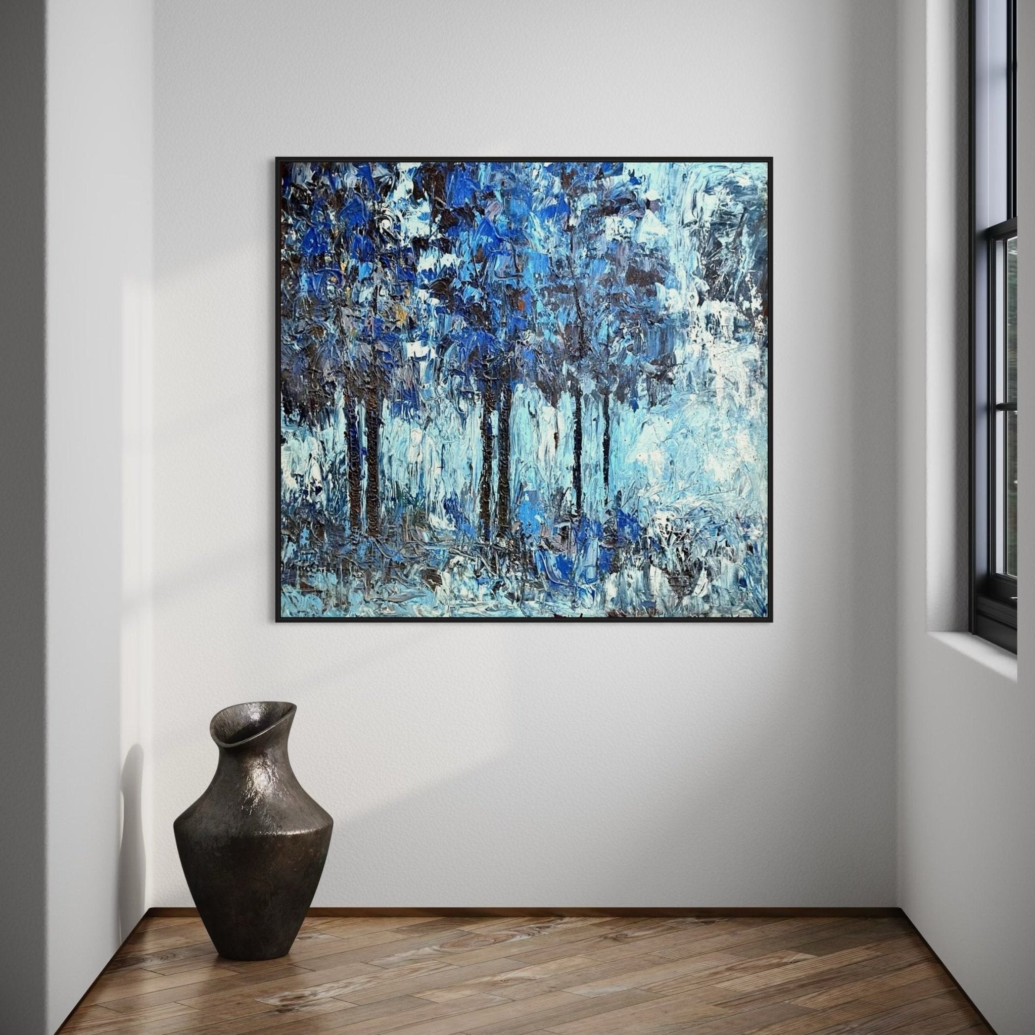 A framed abstract blue and black painting of trees hangs on a white wall above a wooden floor, next to a large window and a dark ceramic vase.