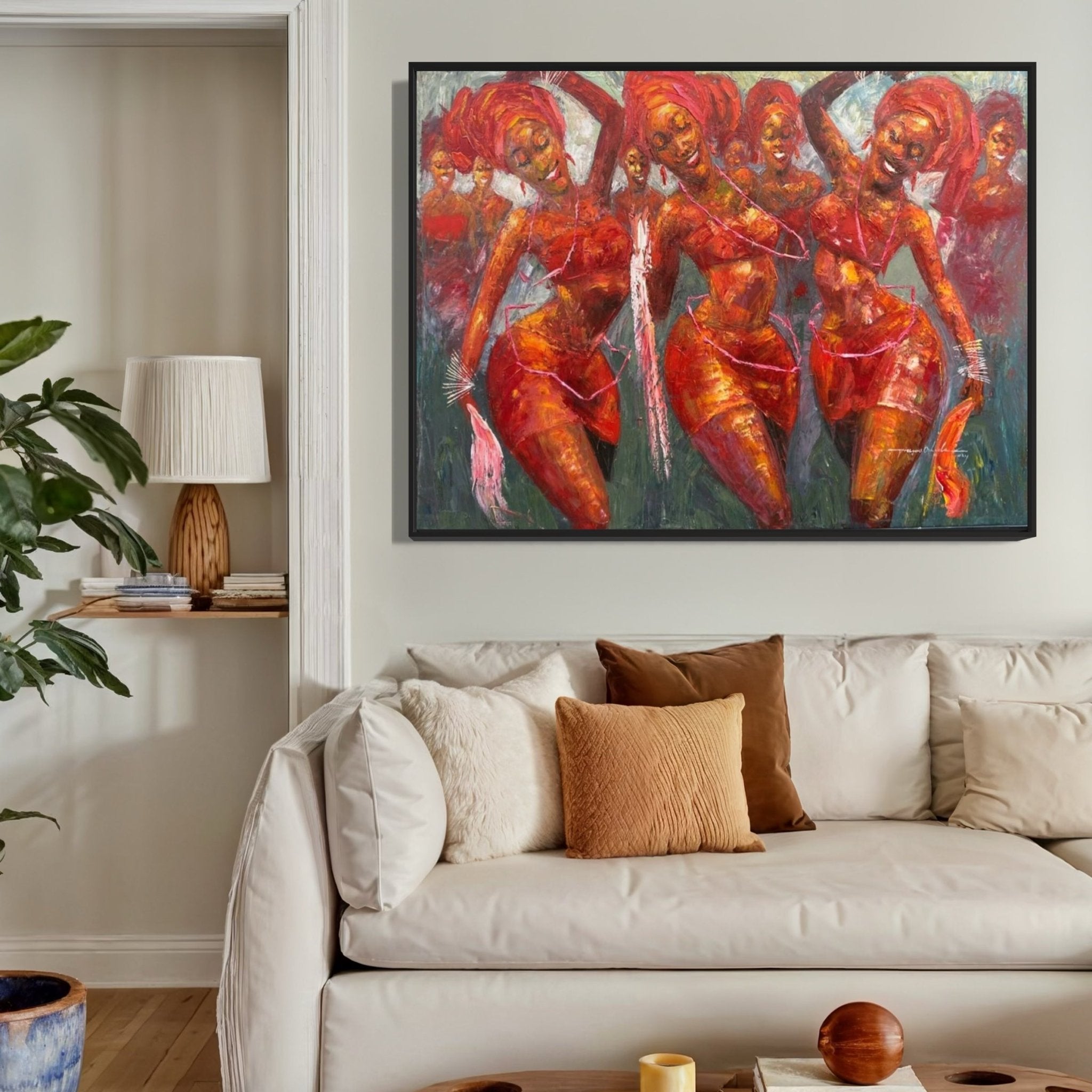 A cozy living room with a light beige sofa, earth-toned pillows, a potted plant, and a colorful painting of dancing women in vibrant red hues hanging on the wall above the couch.