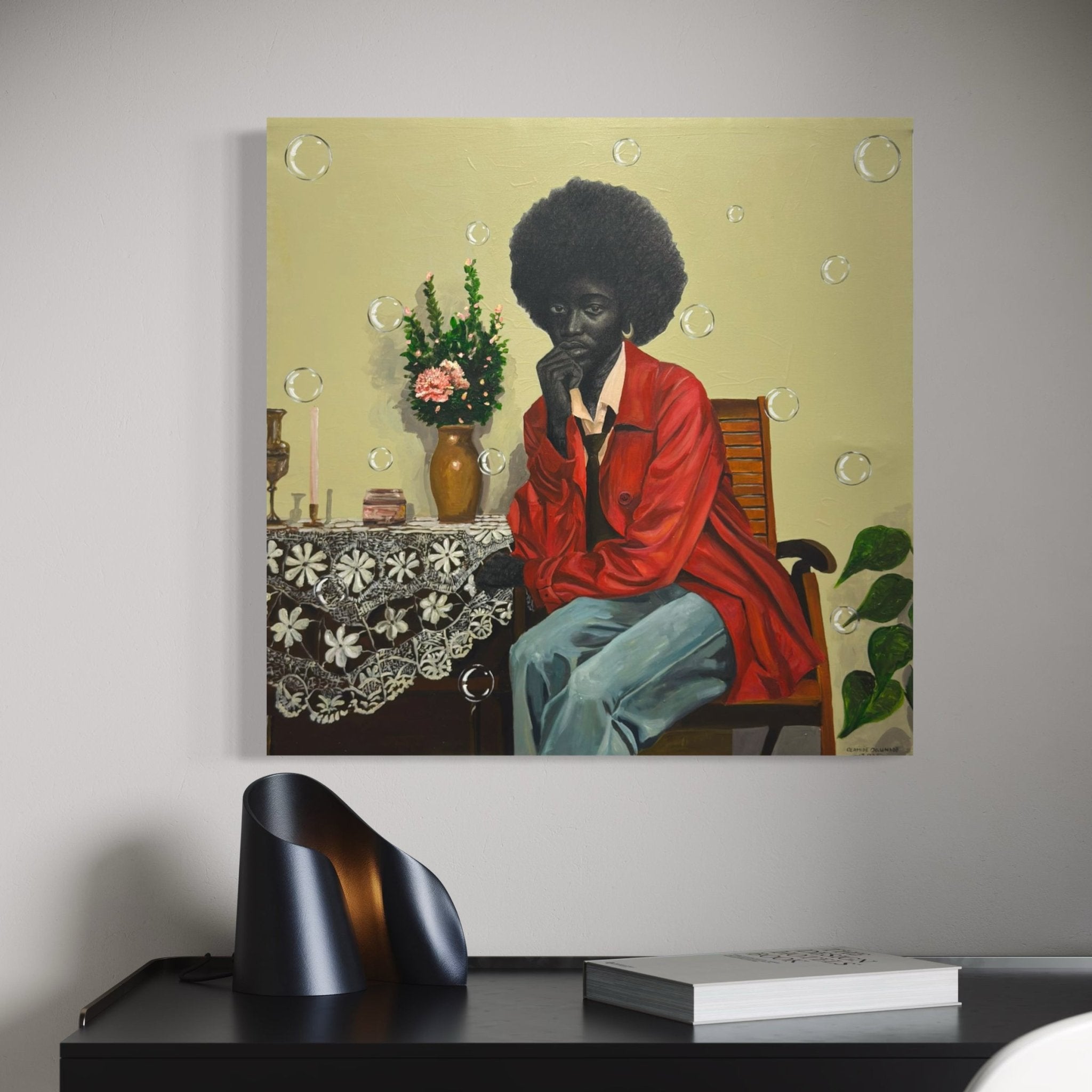 A painting of a person with an afro in a red jacket and blue pants, sitting on a chair by a table with a floral cloth, flowers, and candles, hung on a light wall above a modern black desk.
