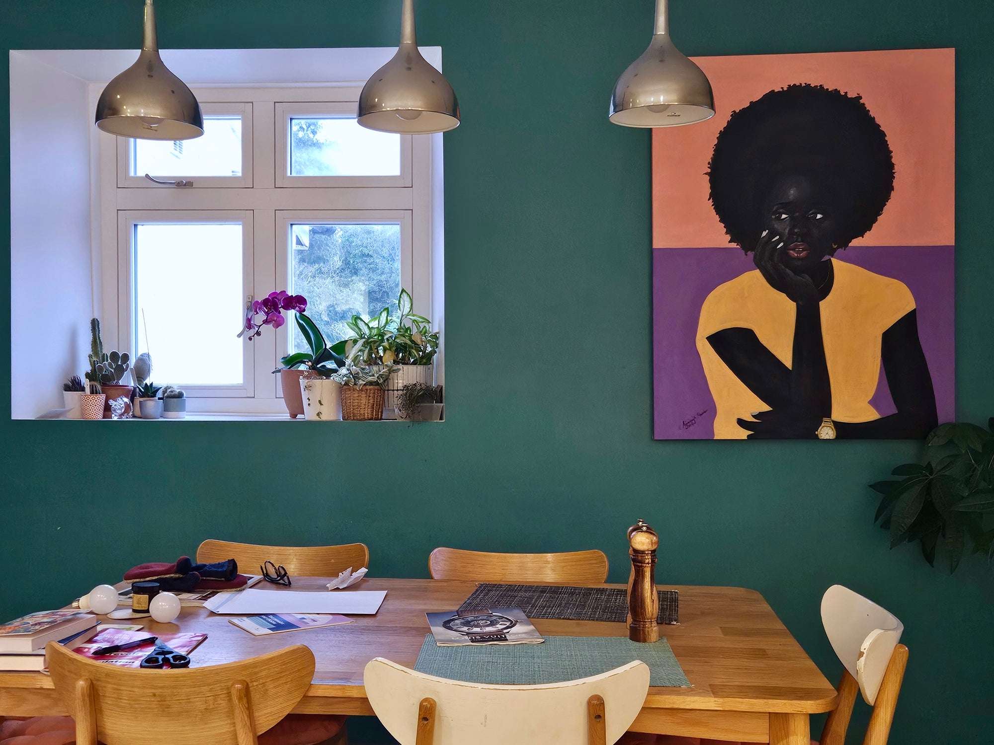 A dining room with green walls, a wooden table and chairs, a window with potted plants, and a large colorful portrait of a person with an afro hanging beside the window. Three pendant lights hang above the table.