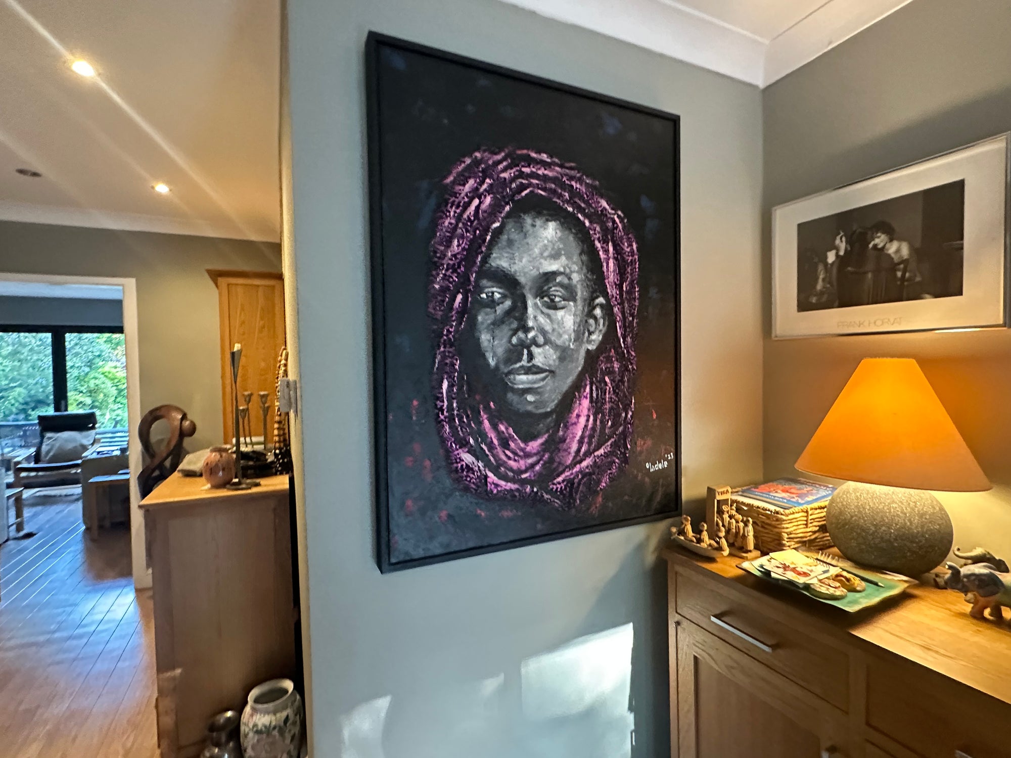 A framed portrait of a person with a pink scarf hangs on a gray wall above a wooden cabinet with a lamp, decorative items, and books in a cozy, well-lit living space.
