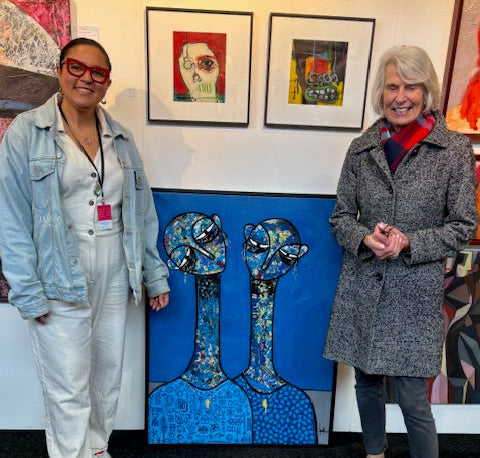 Two women stand smiling in front of colorful artwork at an art exhibition. One wears a light denim outfit and red glasses; the other is in a gray coat. A large blue painting with two abstract figures is between them.
