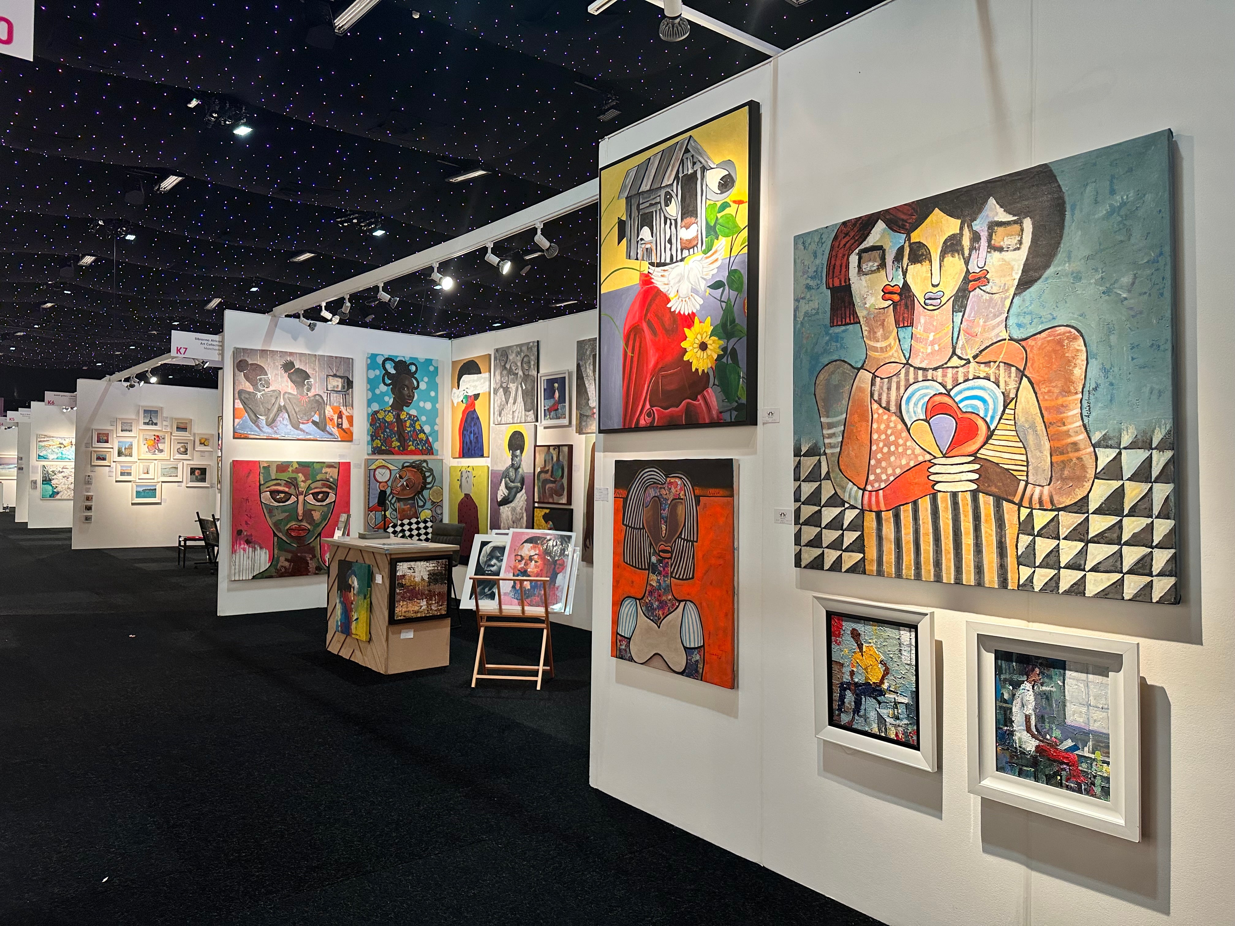 A brightly lit art gallery displays colorful, expressive paintings, mostly portraits and abstract art, on white walls. The gallery has a dark ceiling with star-like lights and paintings arranged neatly along the corridor.