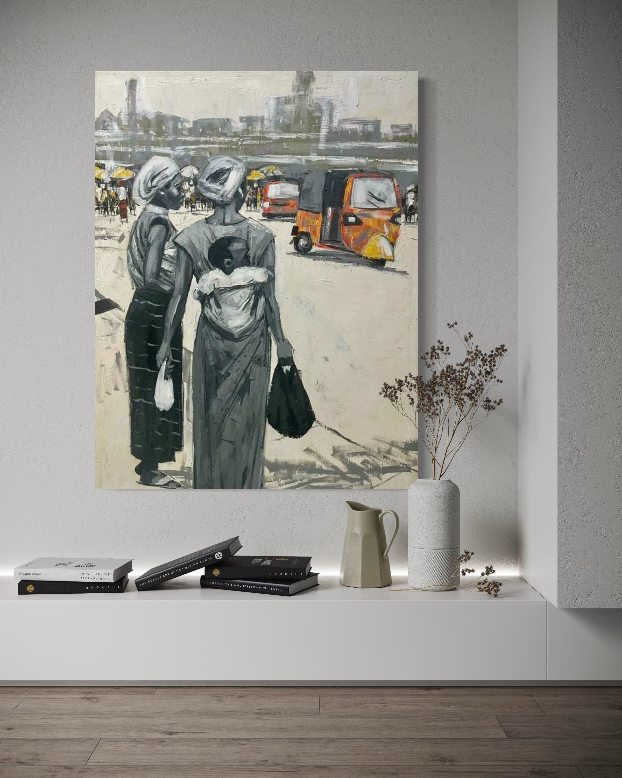 A modern living room showcases 'Lagos Life' City Scape by Lucky Isaiah, a painting of two women in traditional attire with a baby and auto rickshaws—hand-signed by the artist—above books, a vase, and a jug on a white shelf.