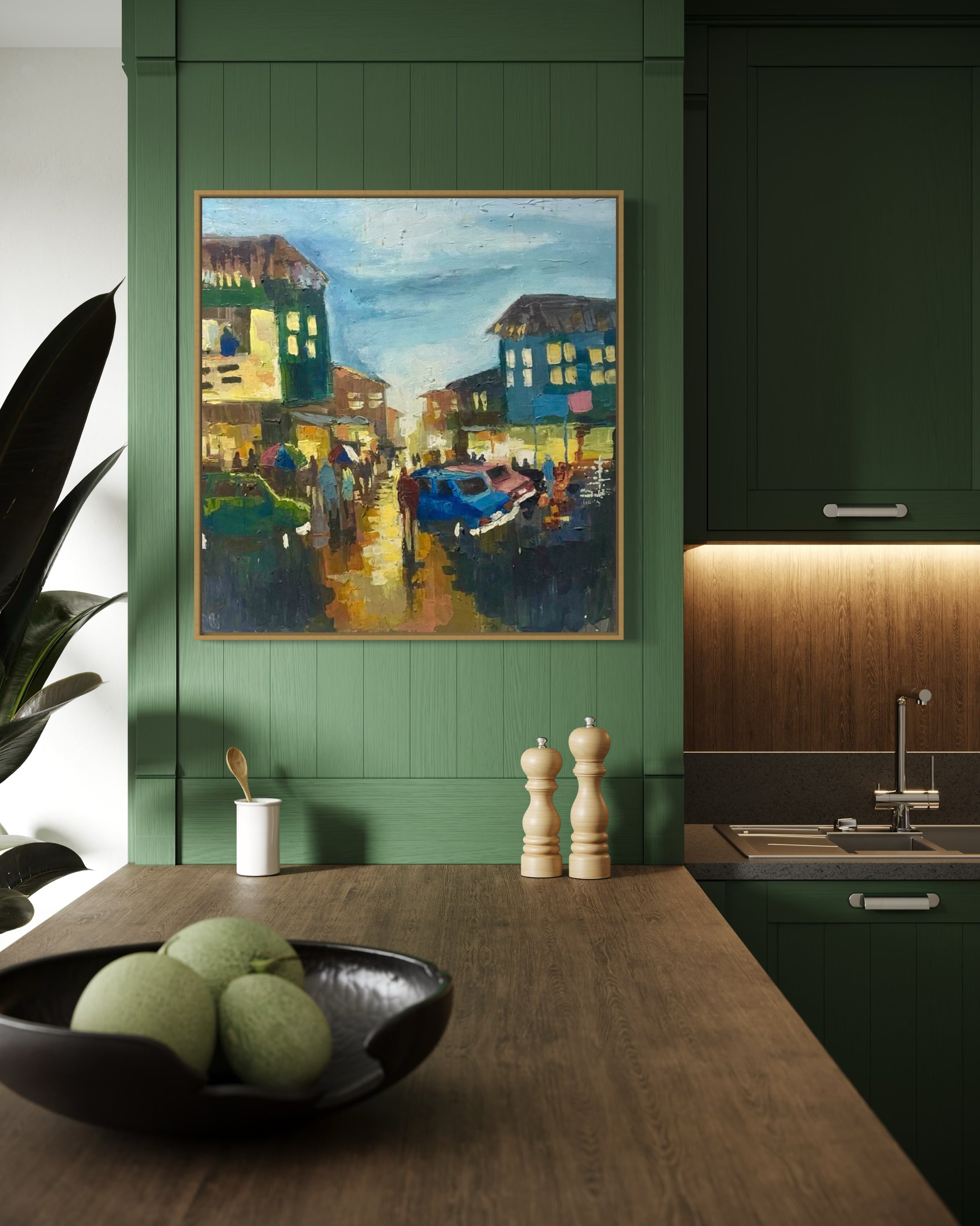 A modern green kitchen with wood accents features 'Somewhere in Ibadan II' by Pitar Alakhume, complete with a certificate of authenticity. On the countertop are two pepper mills, a white cup, and a black bowl filled with green fruit.