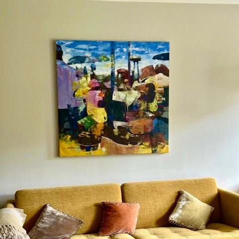 A mustard yellow sofa with assorted cushions sits beneath a colorful abstract painting on a beige wall. A glass coffee table and a folded blanket are also visible in the living room.