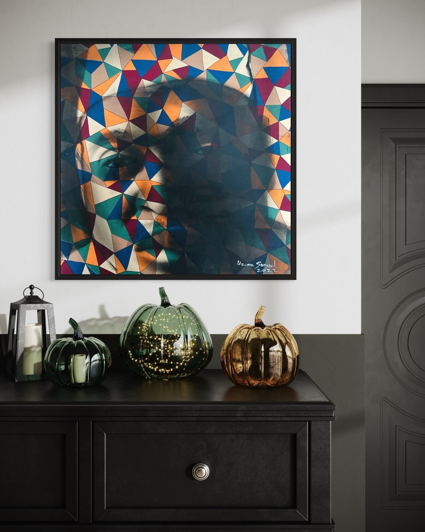 The Abstract Figurative portrait "Self" by Uzoma Samuel hangs above a black dresser accented with three glass pumpkins and lanterns, set against a white and gray wall near a dark, ornate door.