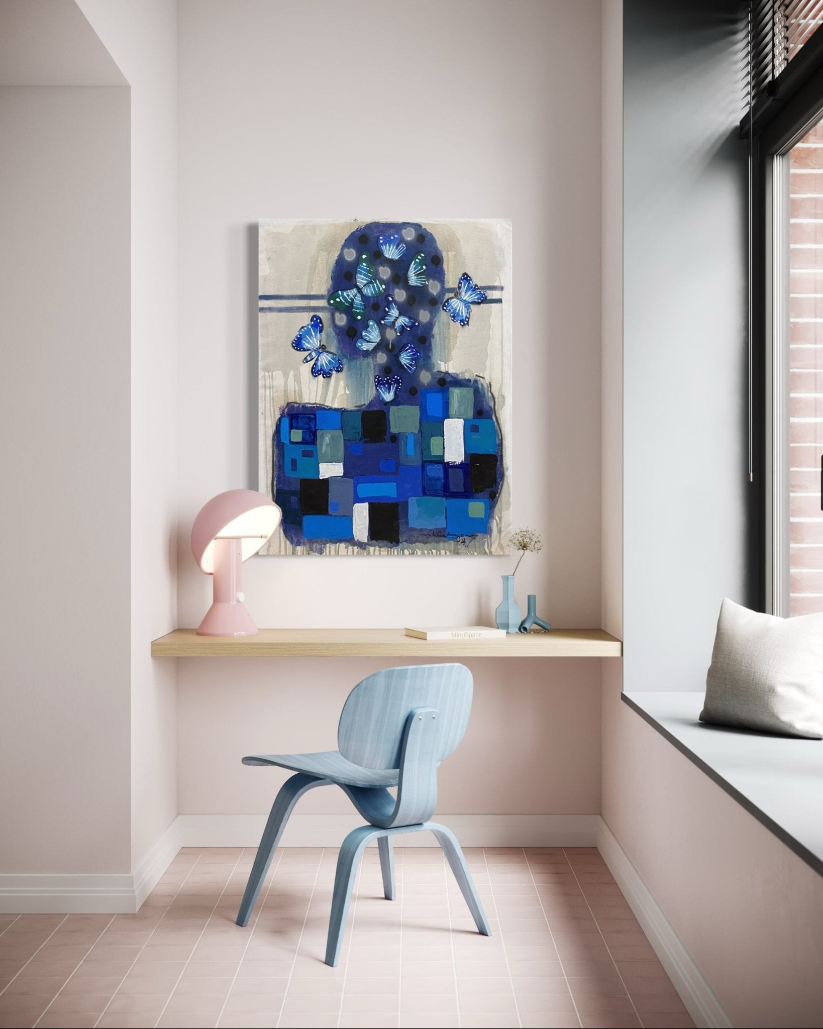 A minimalist workspace features a light blue chair, wood floating desk, pink lamp, small vase, and "Achike Anayo" artwork by Achike Anayo—blue abstract figure with butterfly motifs and certificate of authenticity. Natural light streams in.