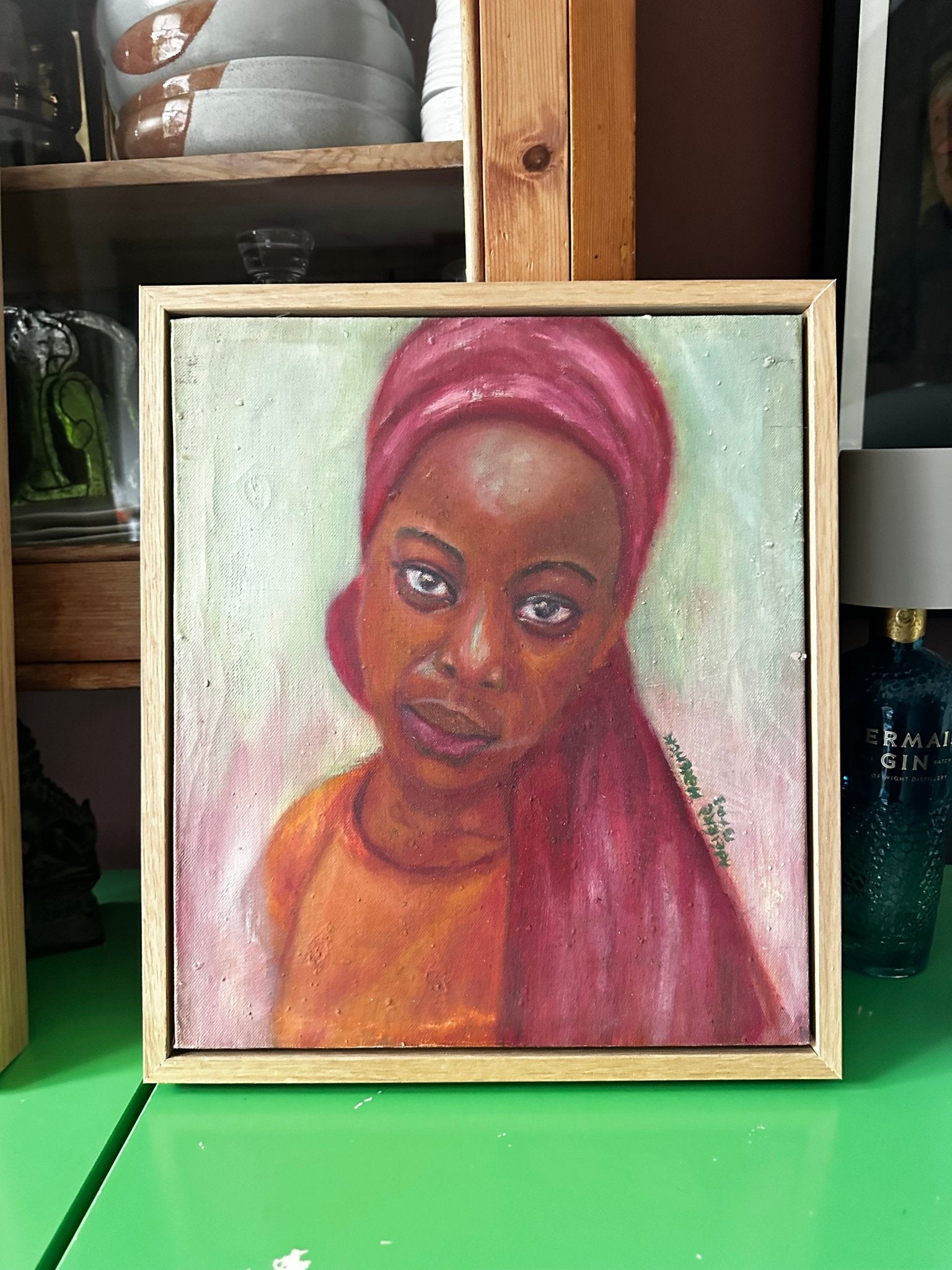 Little Amina" by Bibianna African Art Collective is a hand-signed painting of a woman with a calm expression, in a pink headscarf and orange top, set against green with objects and shelves. Includes certificate of authenticity.
