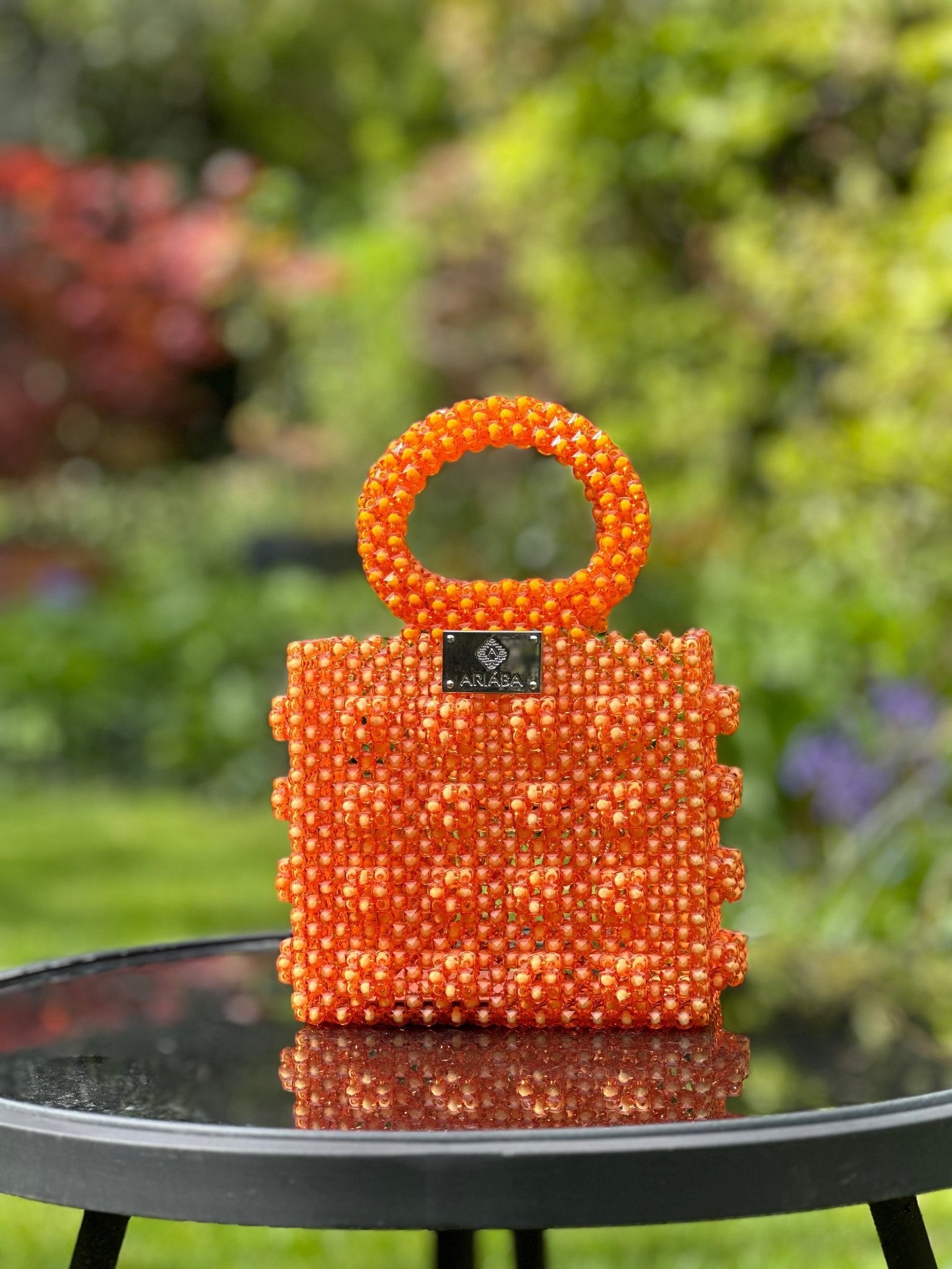 The Ariaba Ira Box Bag Orange by bibianna.co.uk, a handmade designer piece with orange beads and a round handle, rests on a glass table outdoors against a blurred green garden backdrop.