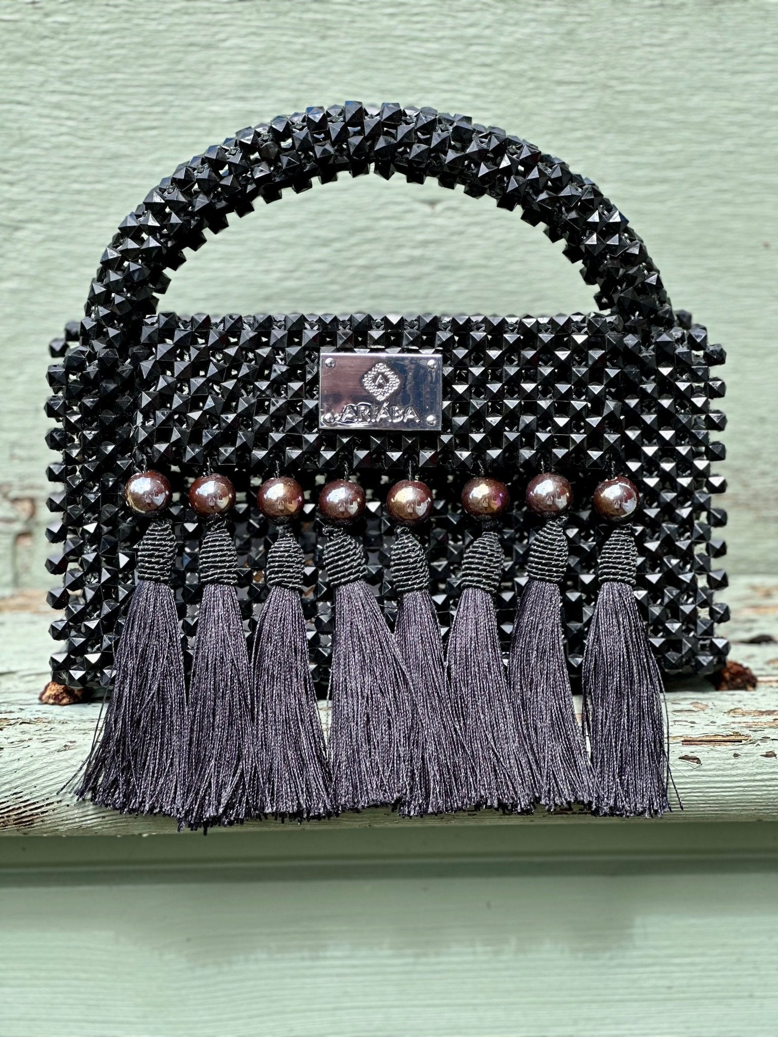 The Ariaba Ira Tasseled Bag - Black by bibianna.co.uk is handcrafted in Nigeria with black beads, metallic studs, and six long tassels on the front, showcased against a pale green background.