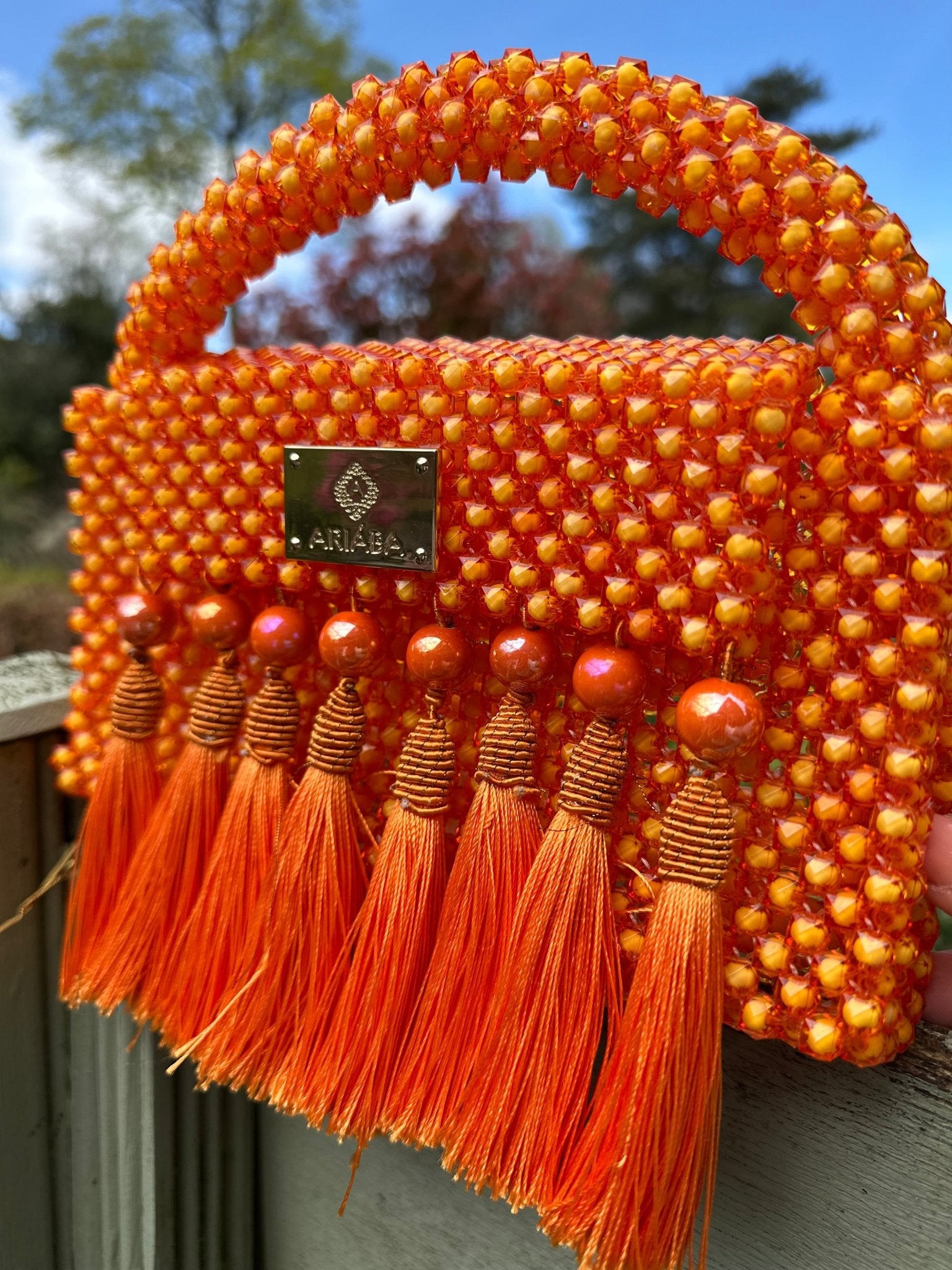 The Ariaba Ira Tasseled Bag - Orange by bibianna.co.uk is a small, handcrafted bag from Nigeria with orange beads, a woven handle, a metallic brand label, and tasseled detailing. It’s shown outdoors against blurred trees.