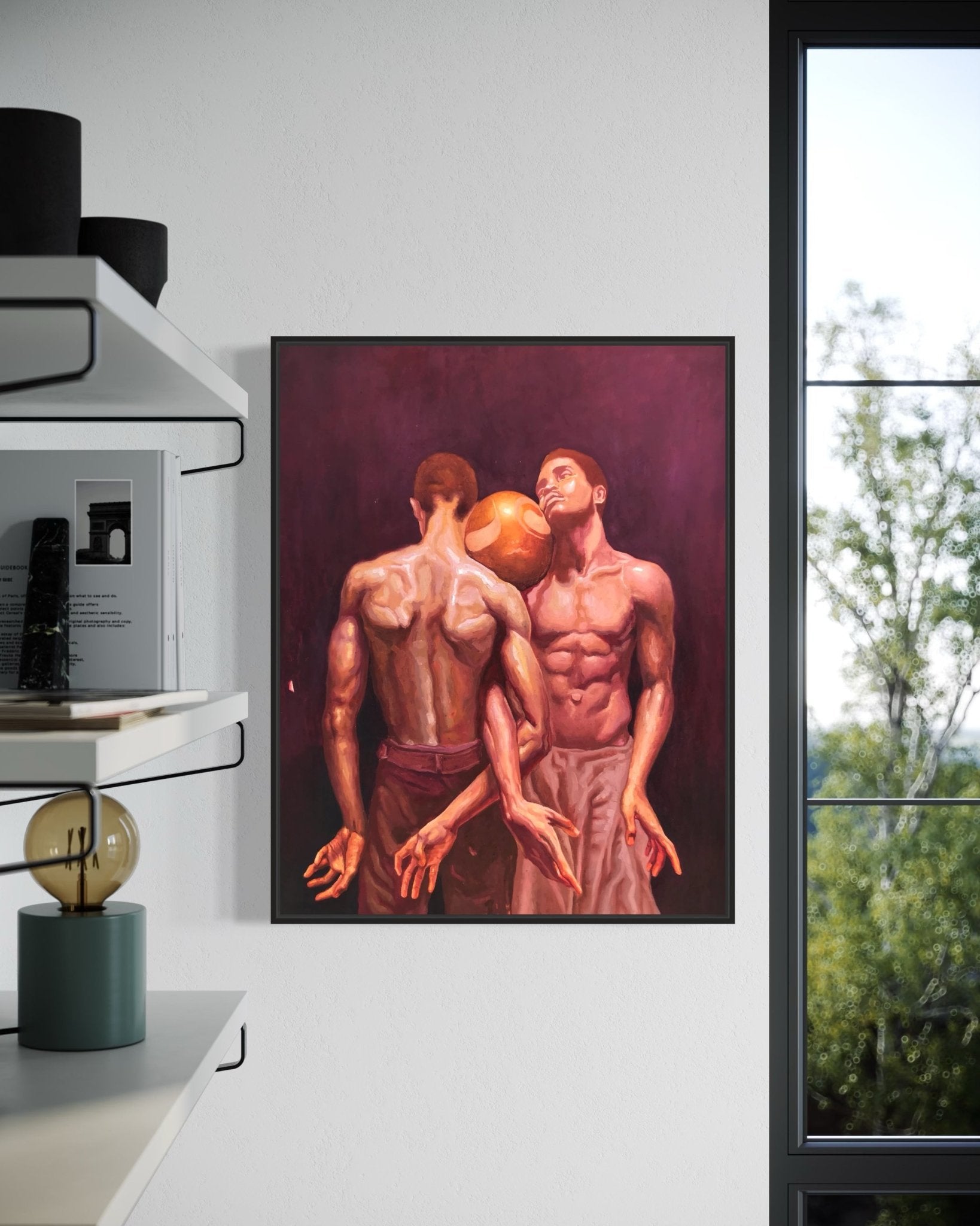 A framed oil painting, "Attached" by Emmanuel Ugwu, depicts two shirtless men with athletic builds balancing a golden ball between their foreheads. The room has white walls, a bookshelf filled with books, and a window facing trees.