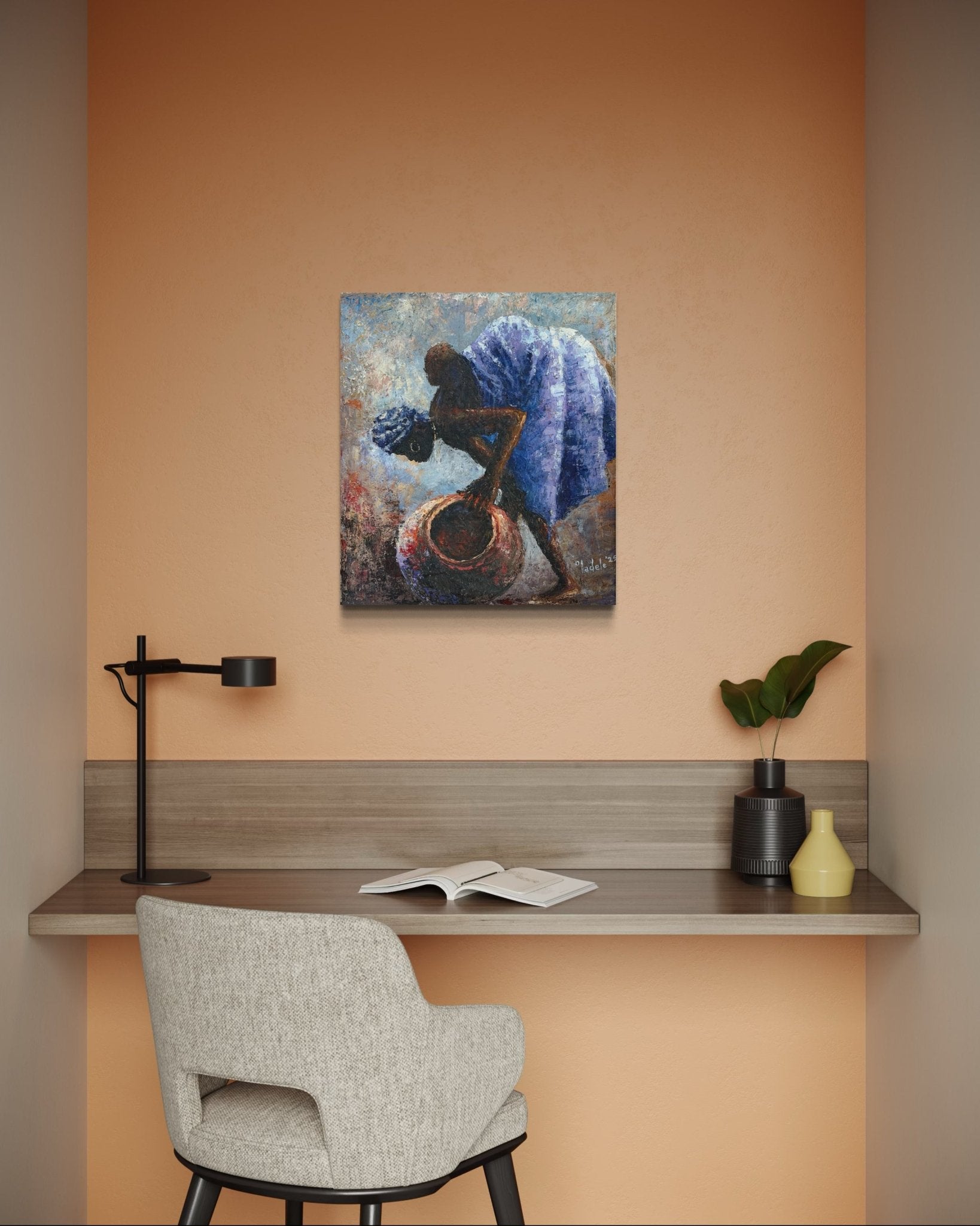A small work desk with a gray chair, black lamp, open book, and vases sits beneath "Backed Up," an original framed African motherhood painting by Sunday Oladele, set against a peach-colored wall.