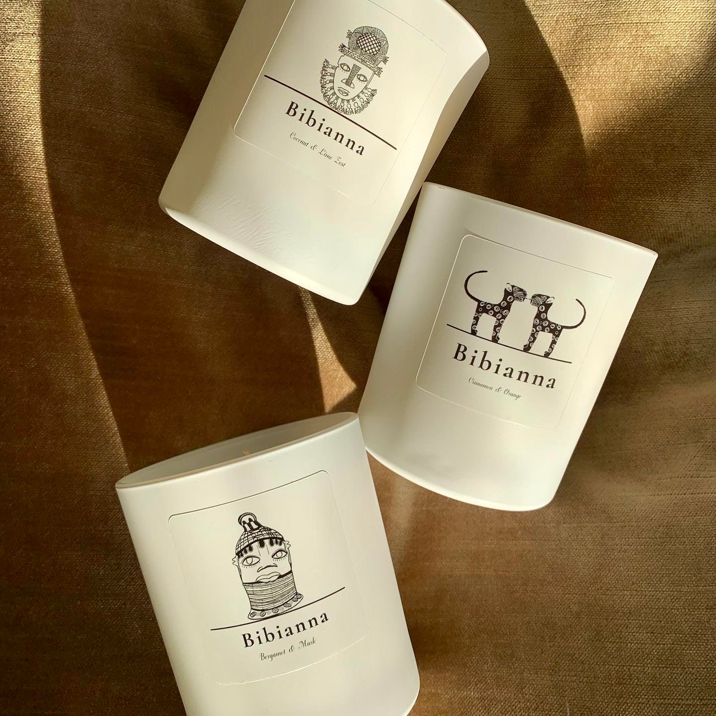 Three white Bibiana candles with minimalist black line art labels and unique face illustrations, arranged on a bronze-colored fabric with warm, natural lighting and soft shadows.
