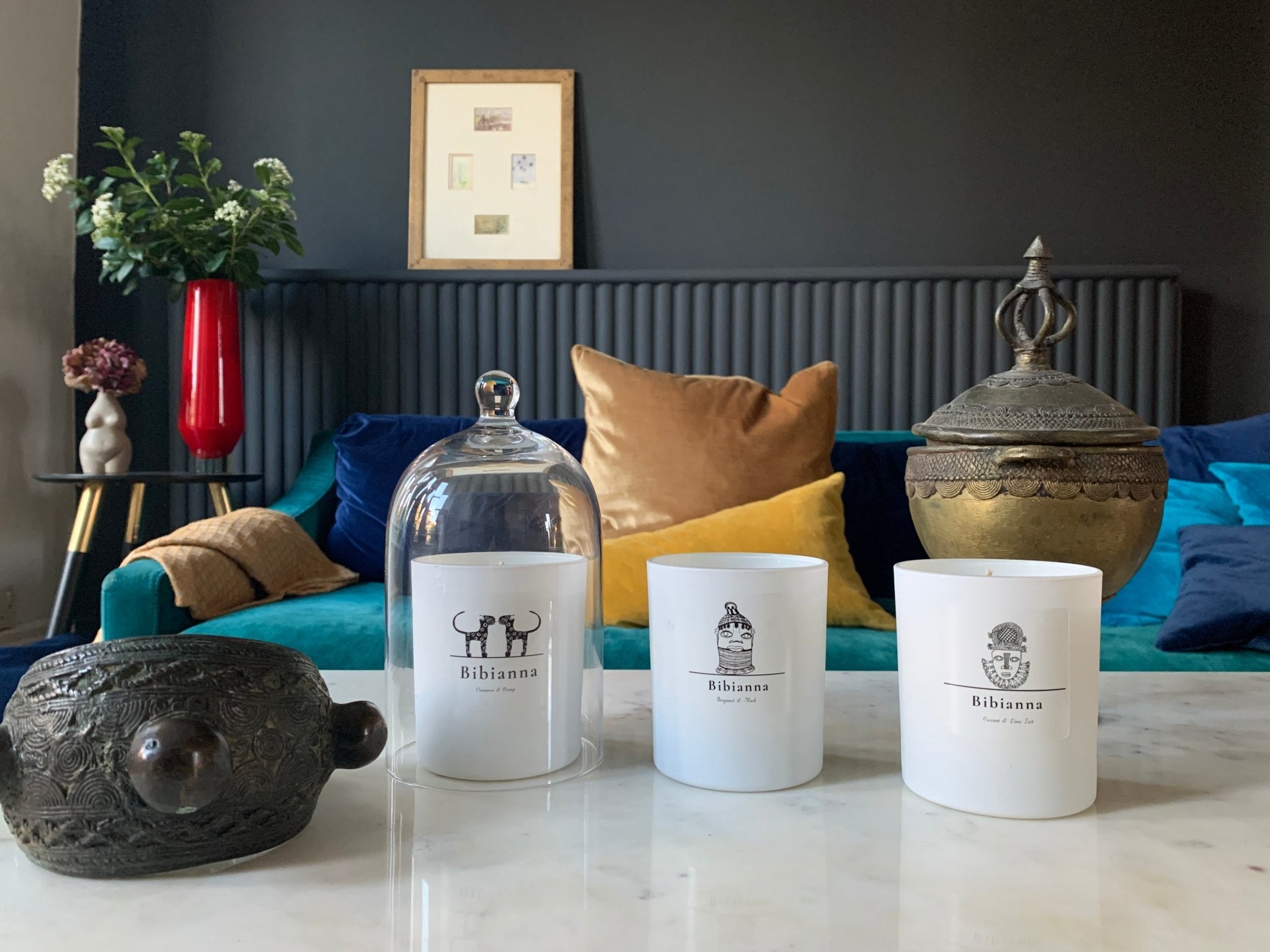 A marble table with three white scented candles, one under a glass cloche, beside ornate metal decor and a turtle figurine. In the background, a blue sofa with colorful cushions and a red vase with flowers.
